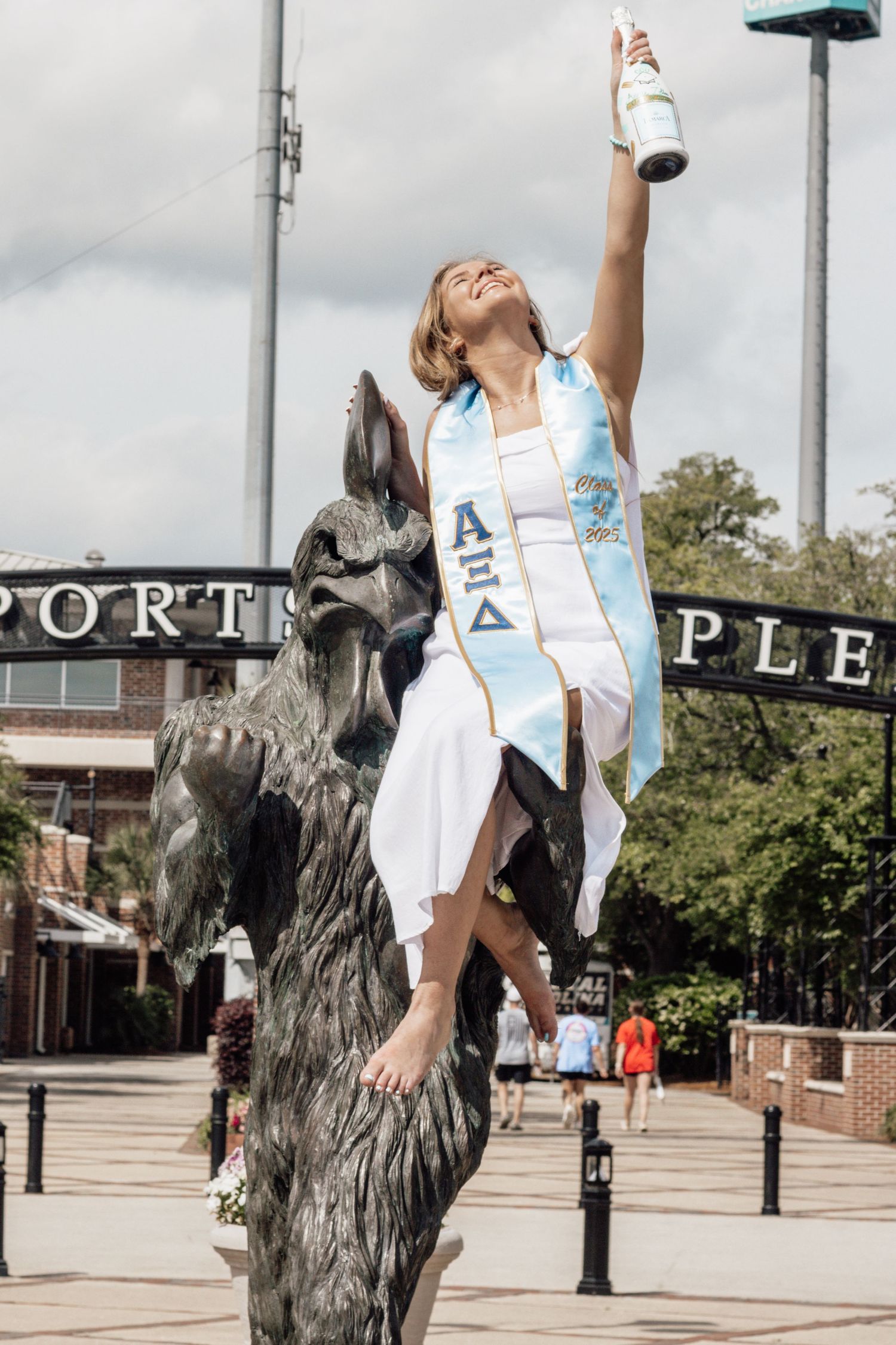 Woman in graduation attire poses playfully next to bronze wolf statues at sports complex.