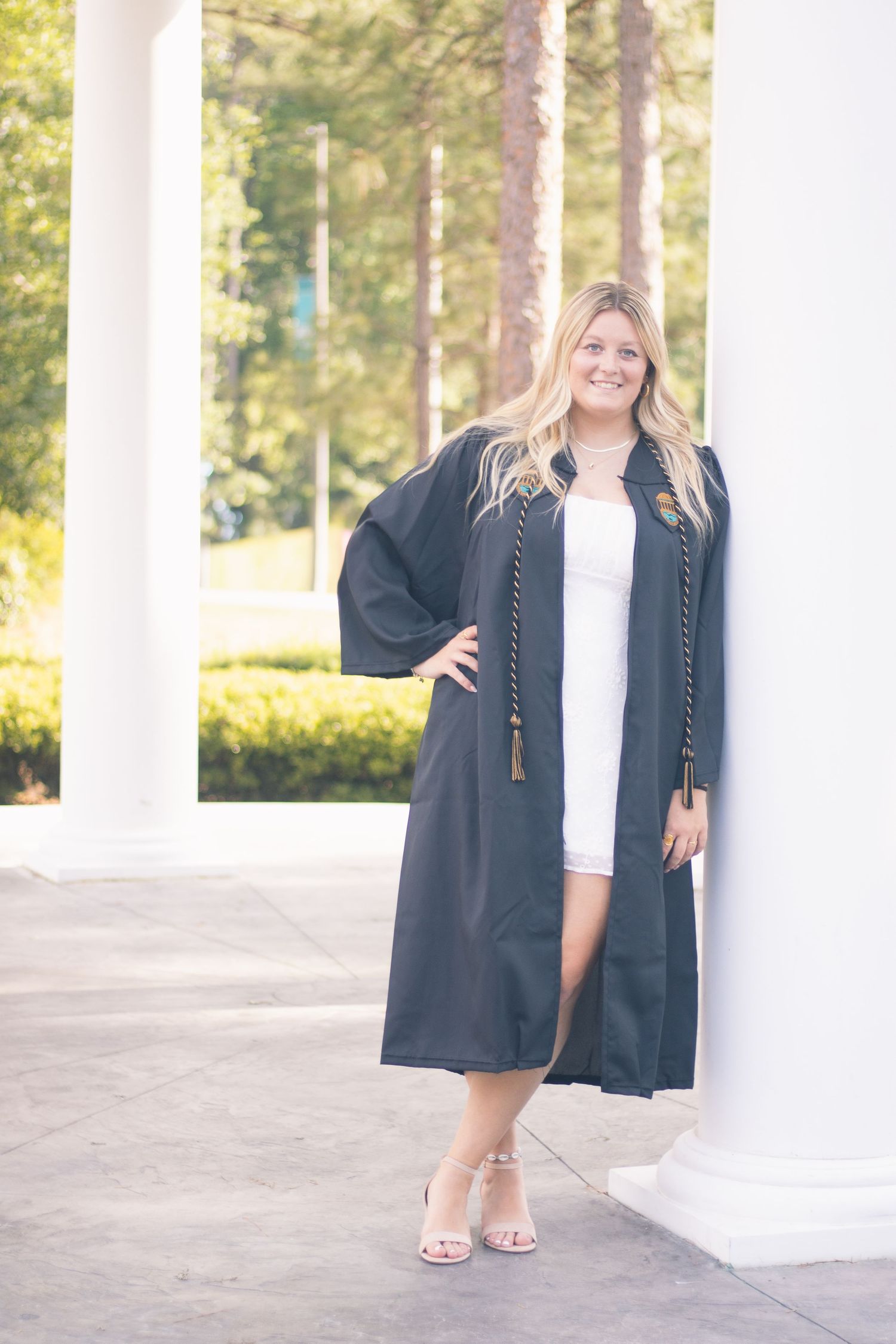Graduate in black gown poses between white columns in soft natural lighting.