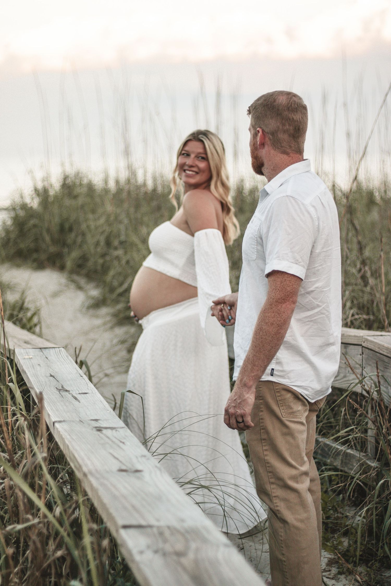 Couple posing together on wooden boardwalk with coastal dunes in background.