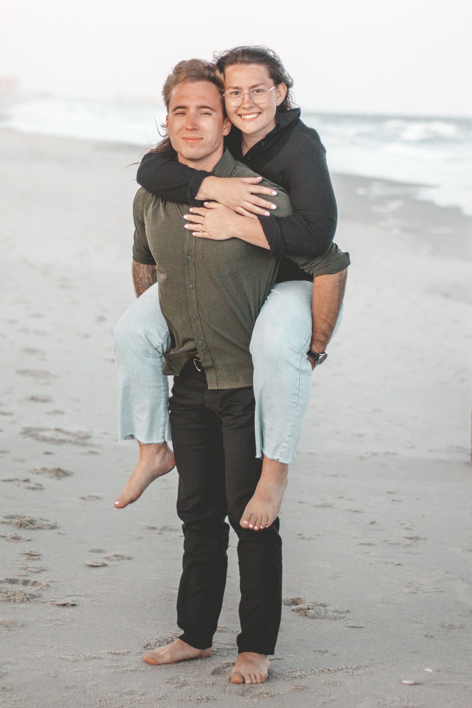 Two people share a playful piggyback ride moment on the beach with ocean waves in the background.