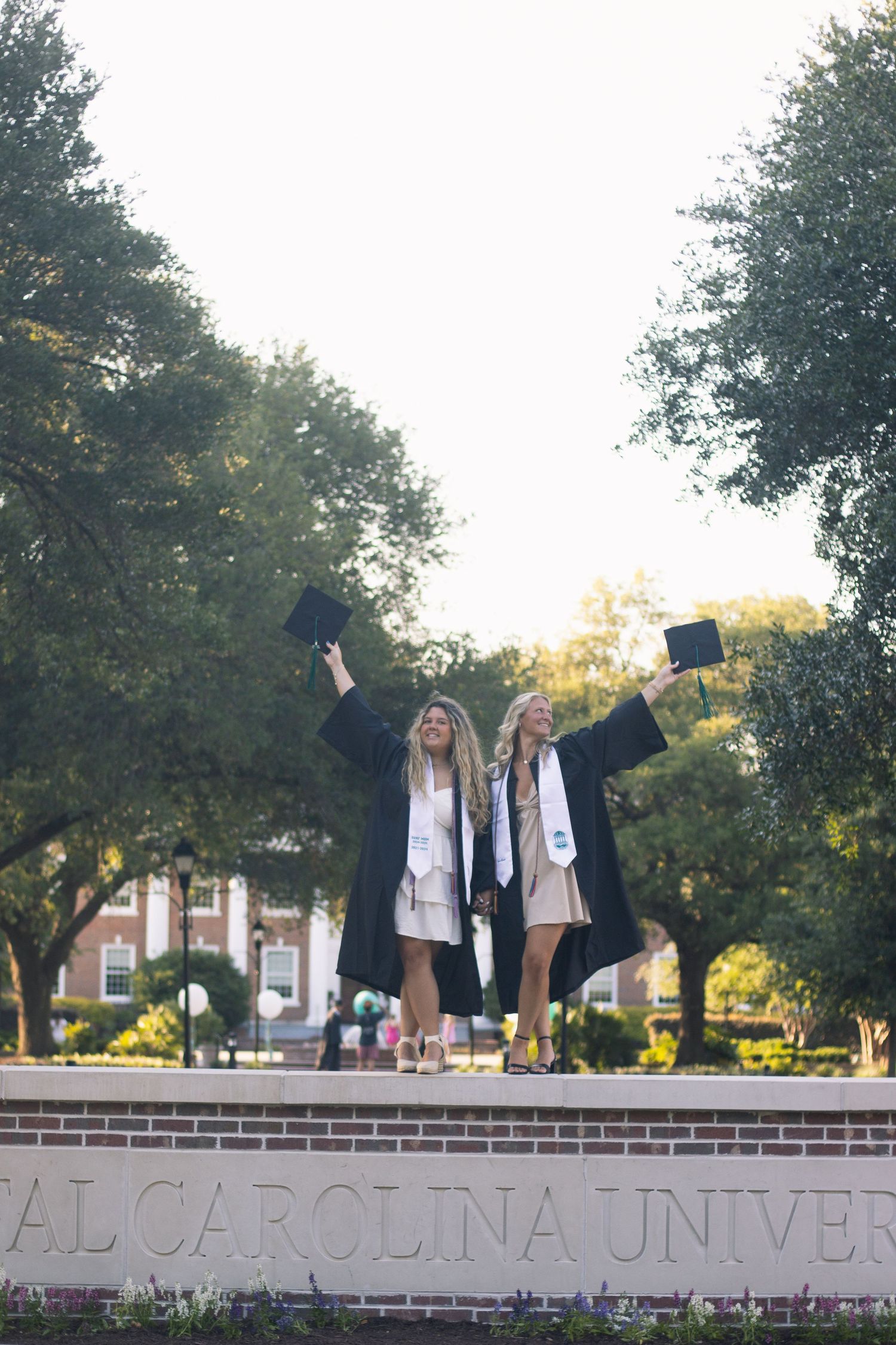 Graduates celebrate on university sign wall while holding caps in the air.