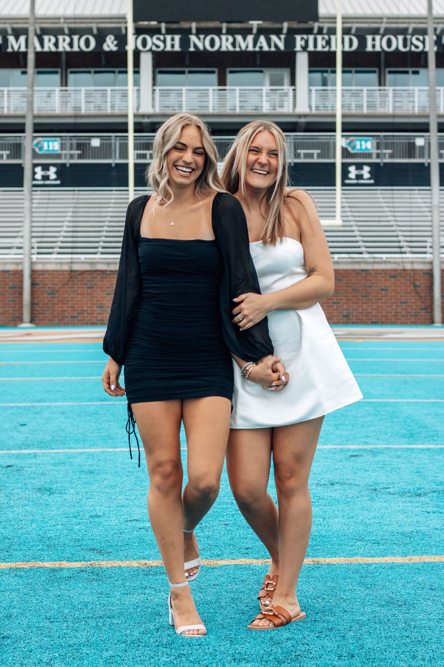 Two graduates pose on blue turf field in front of stadium scoreboard.