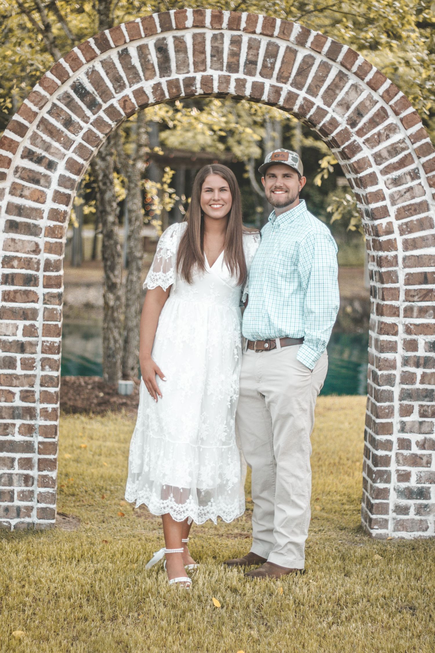 A couple stands beneath a decorative brick archway in classic white and casual attire.