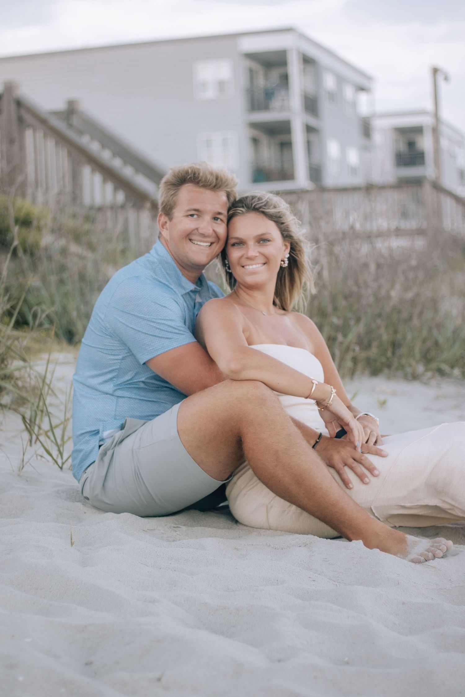 Couple sitting together on sandy beach with apartment building in background.