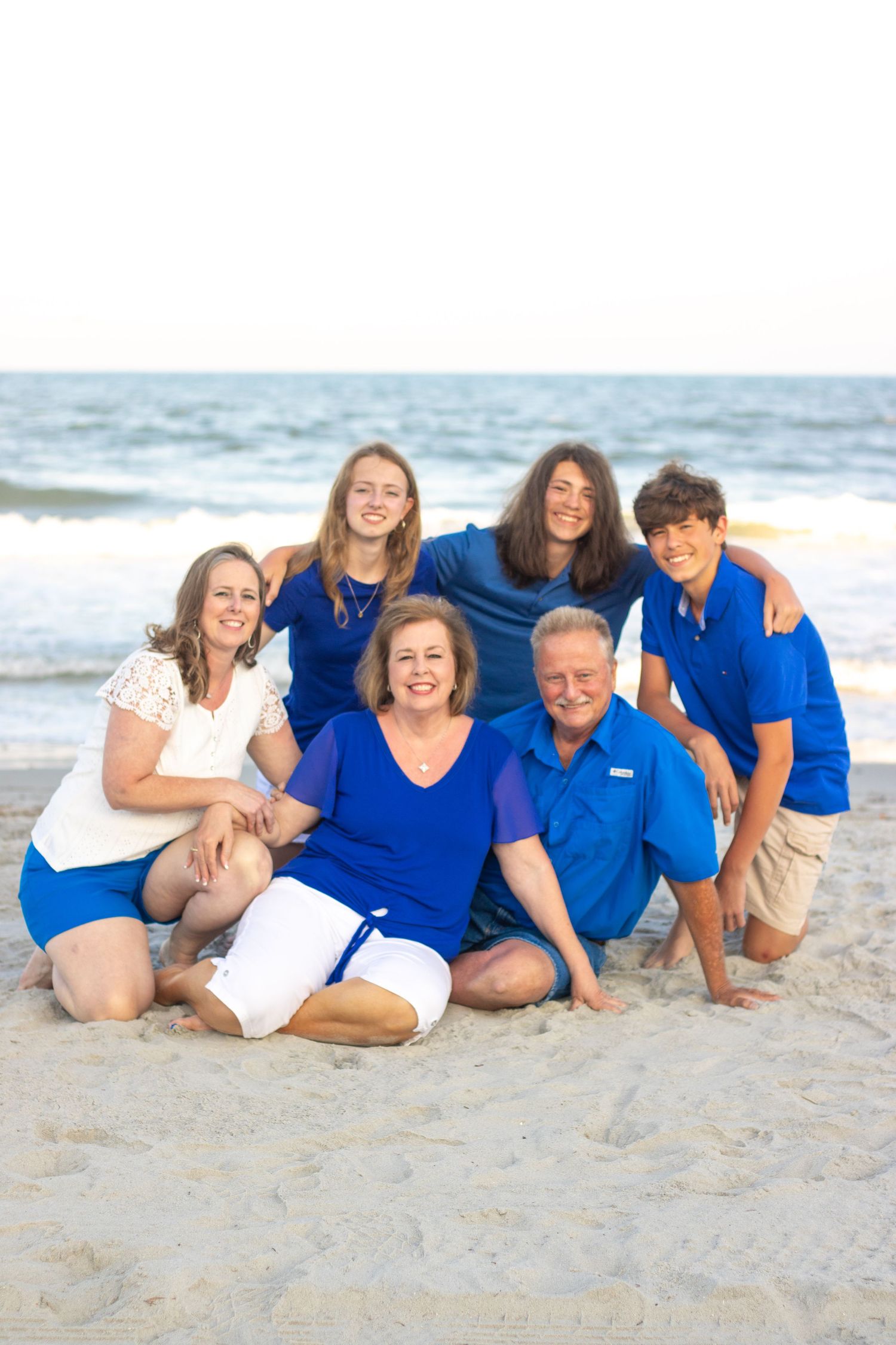 Extended family group wearing coordinated blue outfits poses together on sandy beach at sunset.