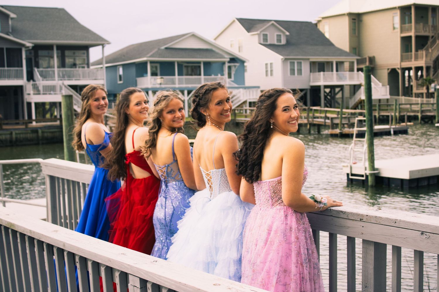 Wedding party members in colorful dresses standing on a waterfront deck near coastal homes.