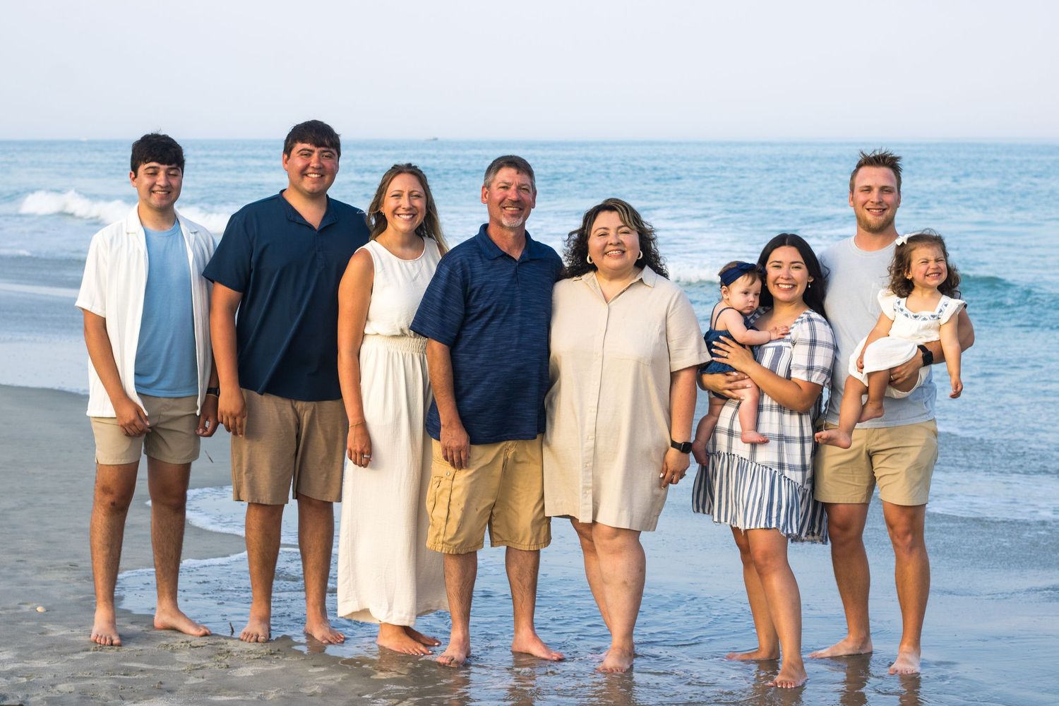 Large family group wearing coordinated white and navy outfits poses along oceanfront at dusk.