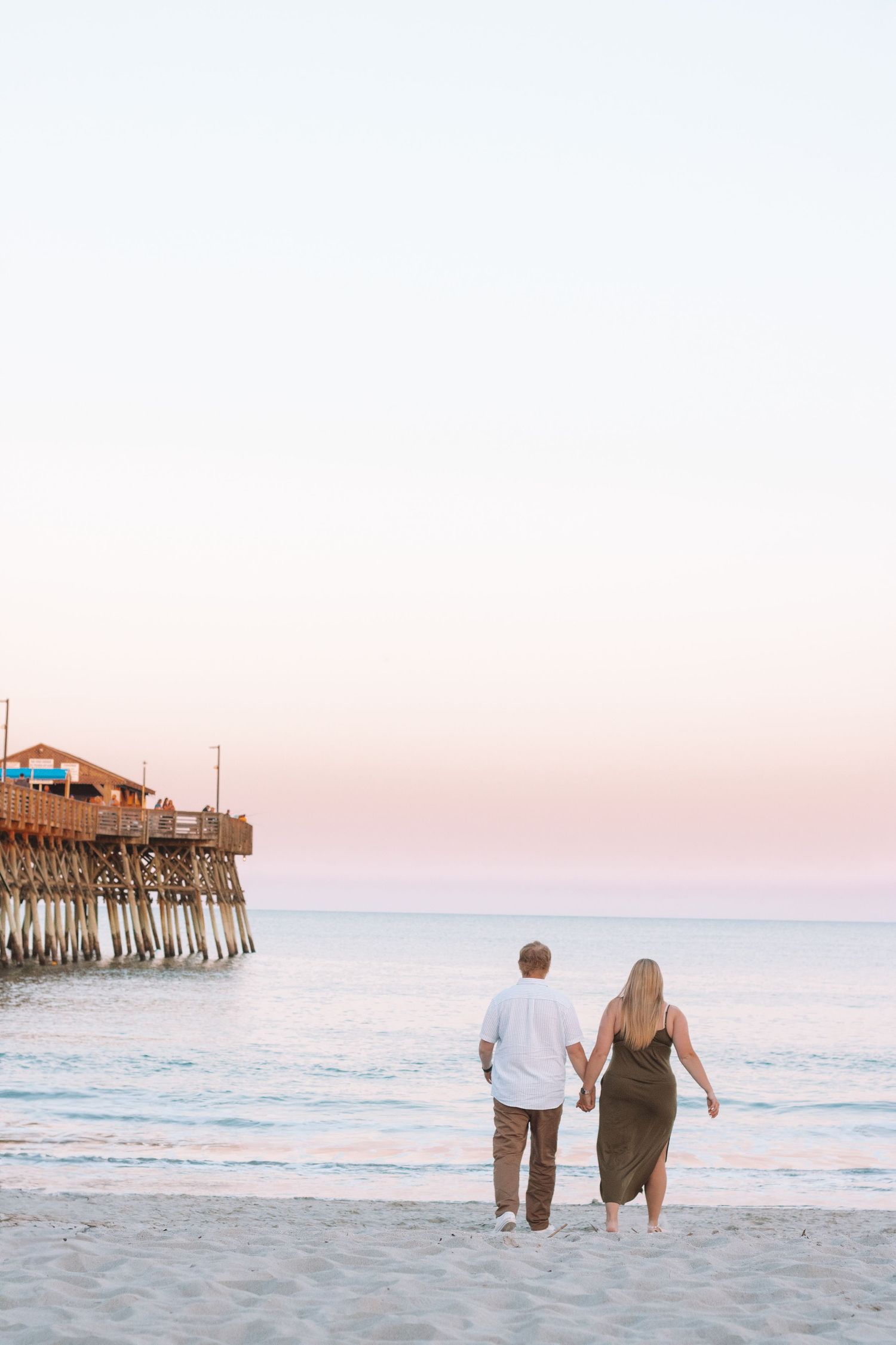 A couple walks hand in hand on the beach at sunset near a wooden pier.