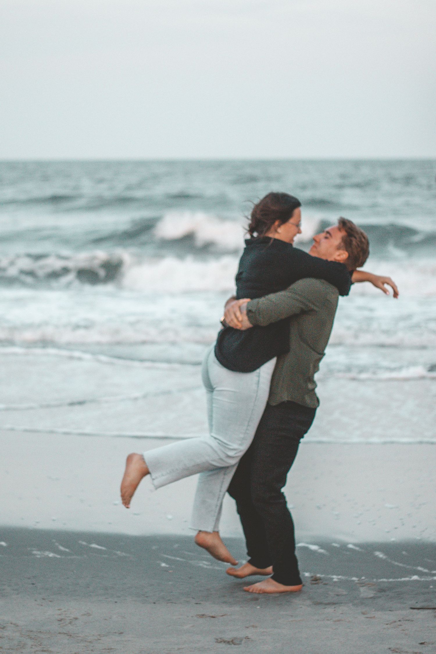 A couple shares a joyful embrace on the beach with crashing waves behind them.
