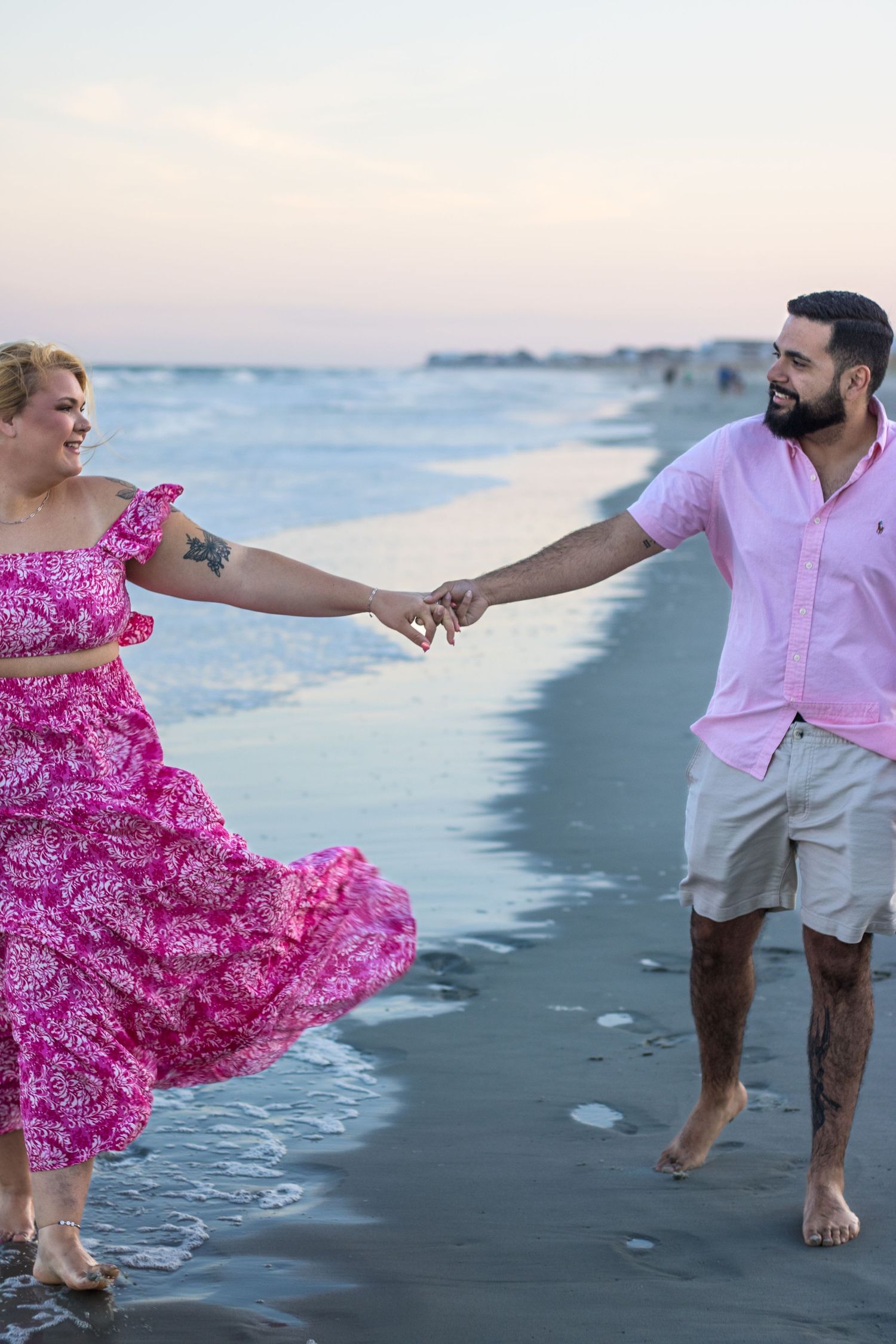 Couple walking barefoot along beach shore at sunset wearing pink and white outfits with flowing dress.