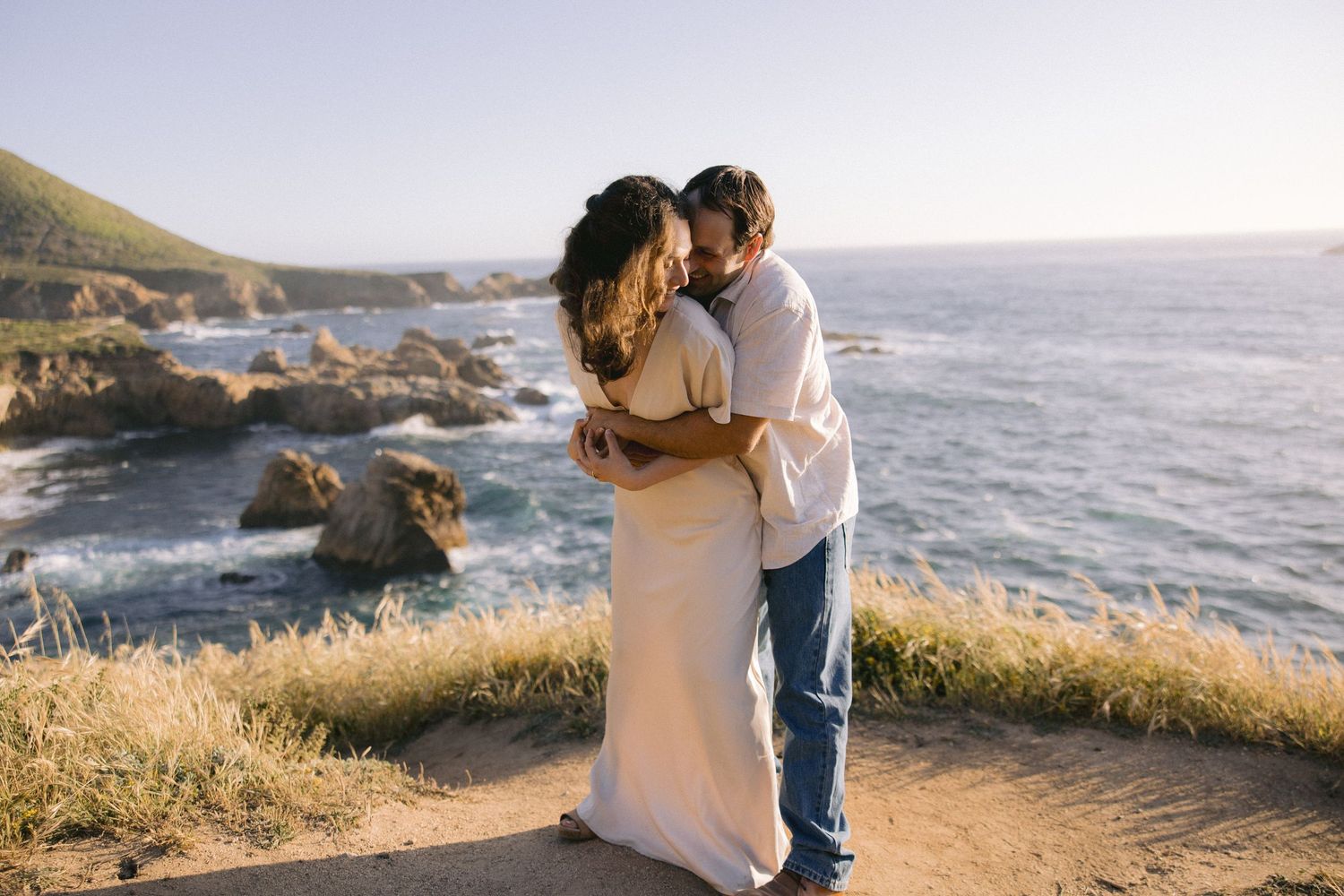 Couple embraces in romantic moment at scenic coastal cliff overlooking ocean at sunset.