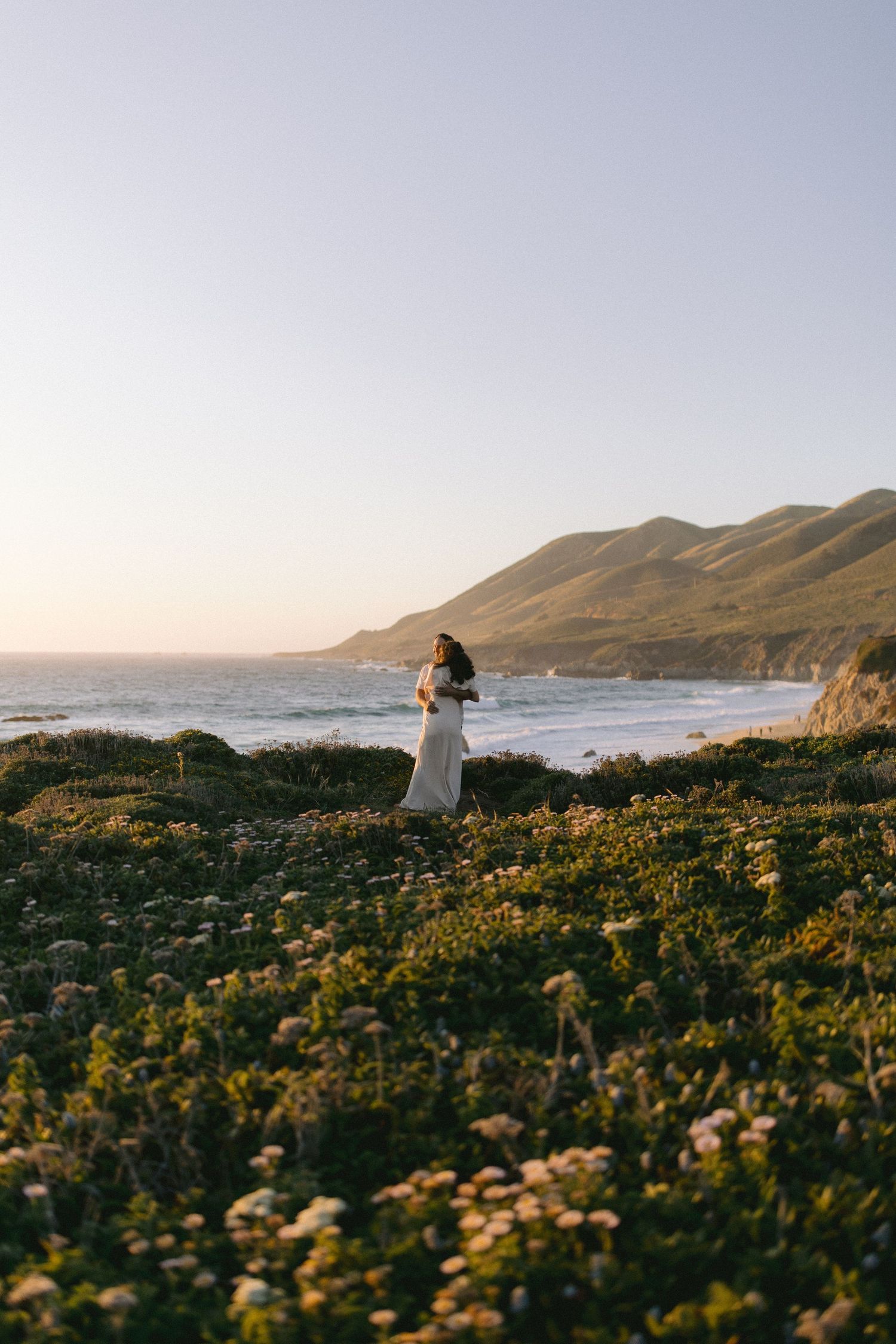 Figure in white dress stands on coastal cliff overlooking Big Sur coastline with wildflowers at sunset.
