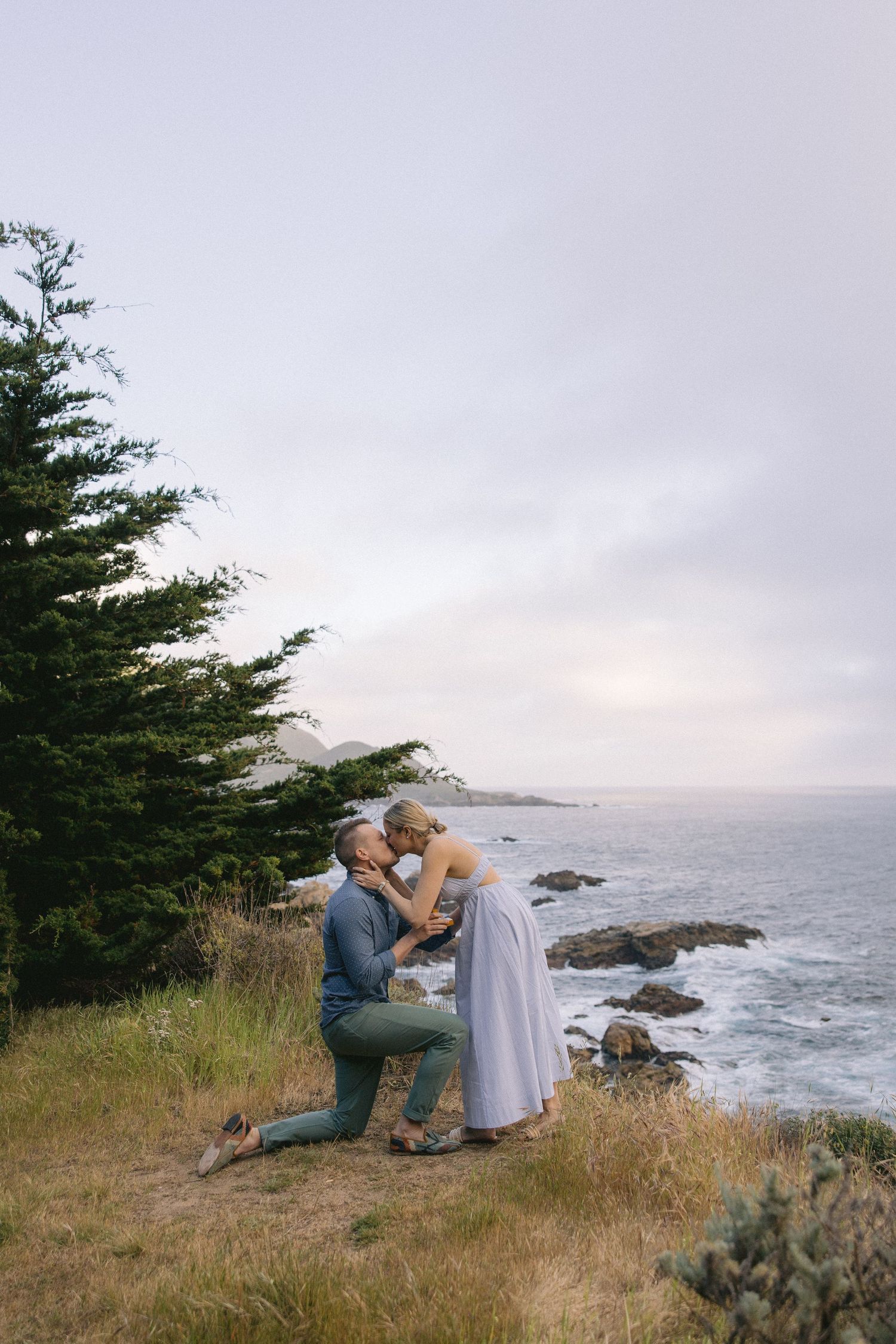 A tender moment shared between a couple on a cliff overlooking a rocky coastline with evergreen trees at sunset.