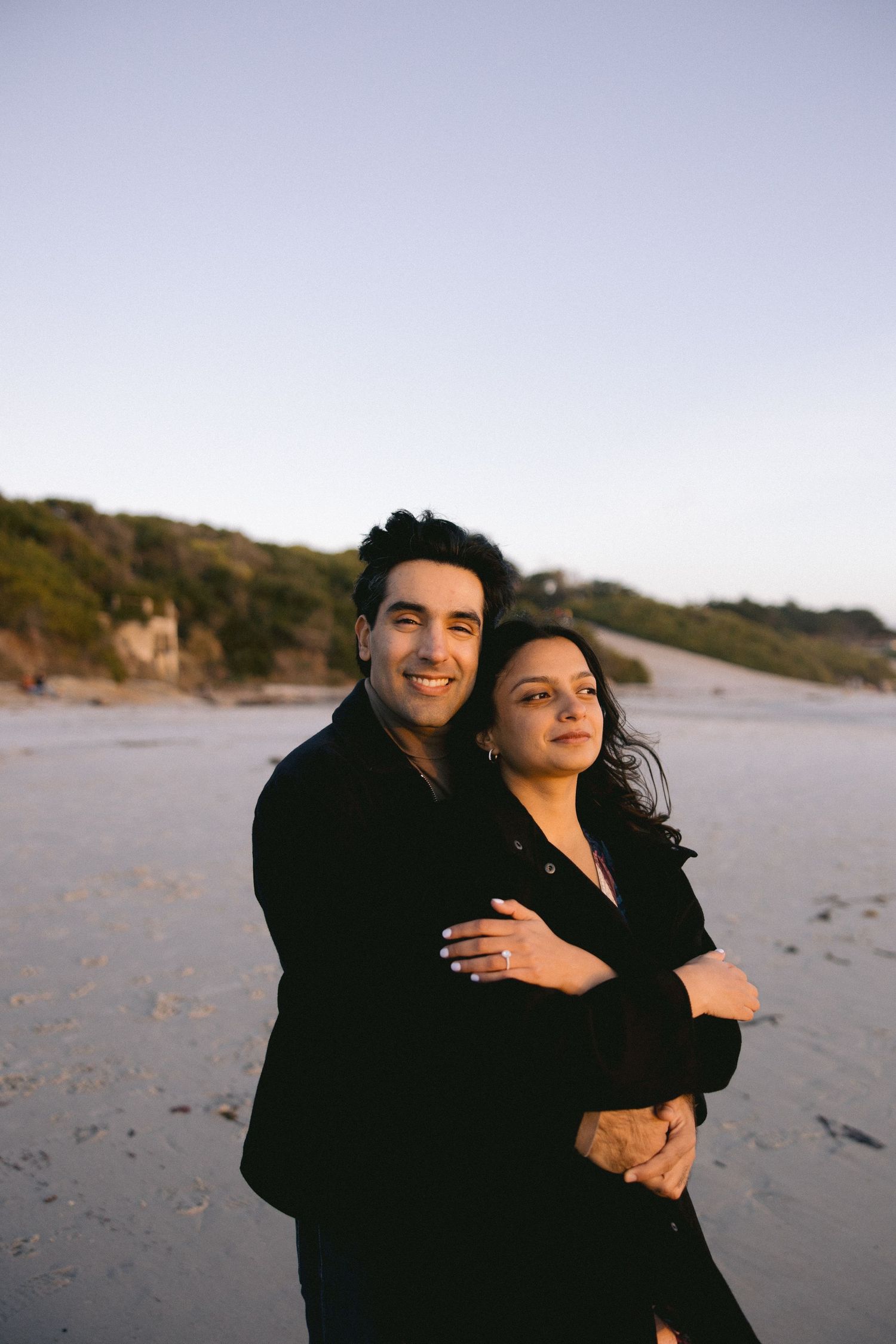 A couple embraces on a beach at dusk wearing matching black outfits with hills in the background.