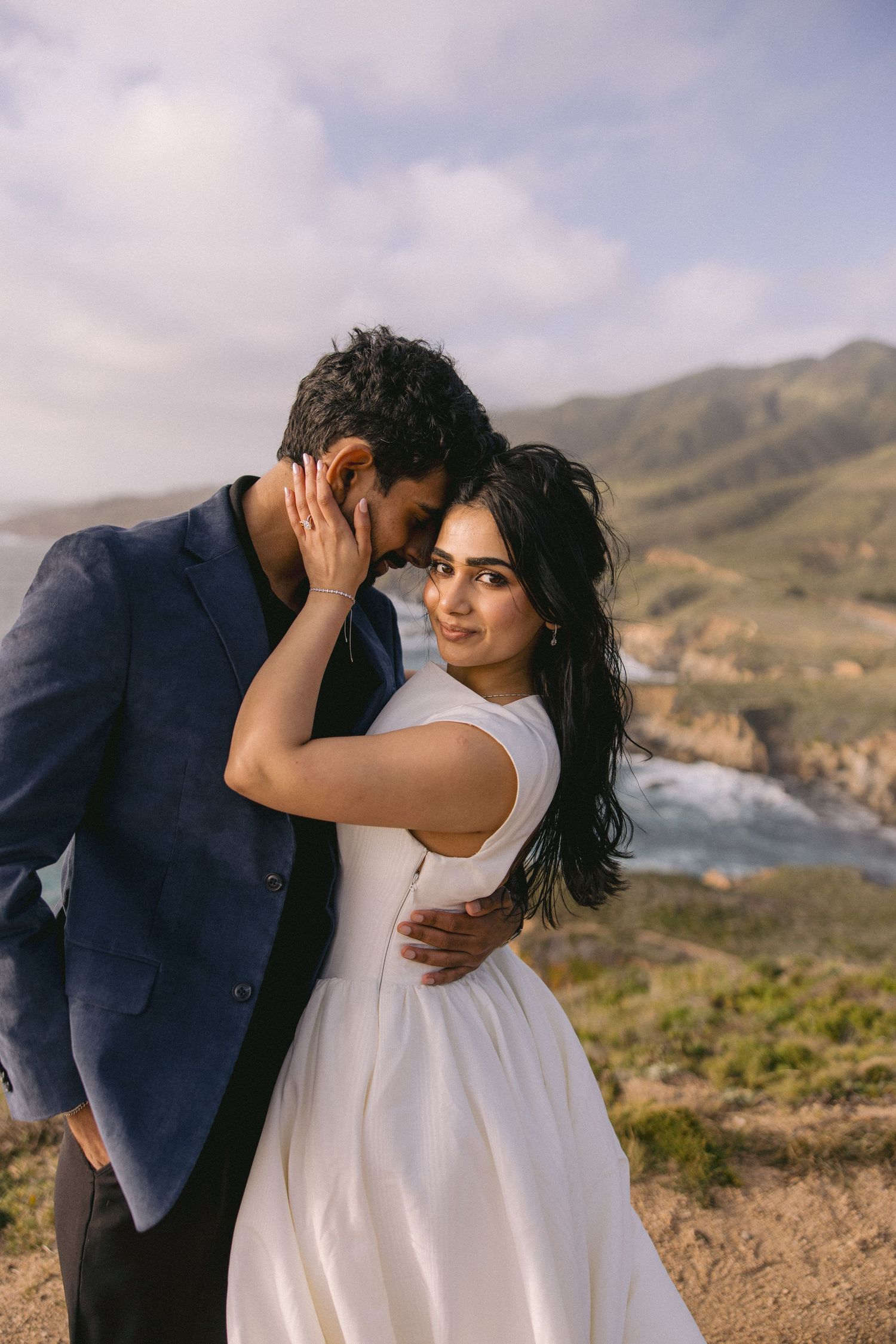 A romantic couple embraces on a coastal clifftop overlooking rugged mountains and ocean on their wedding day.