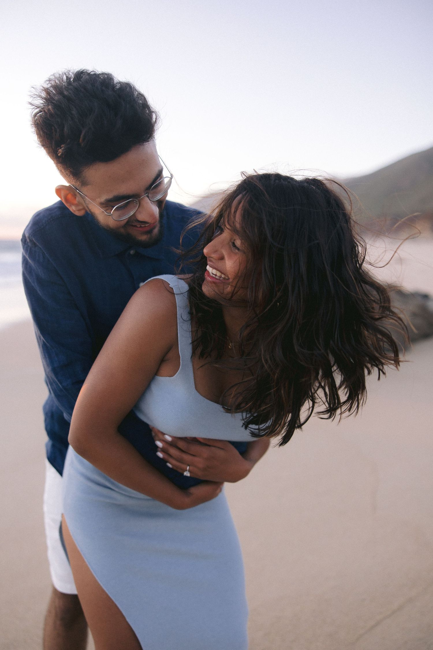 A romantic couple embraces and laughs together on a beach at sunset.
