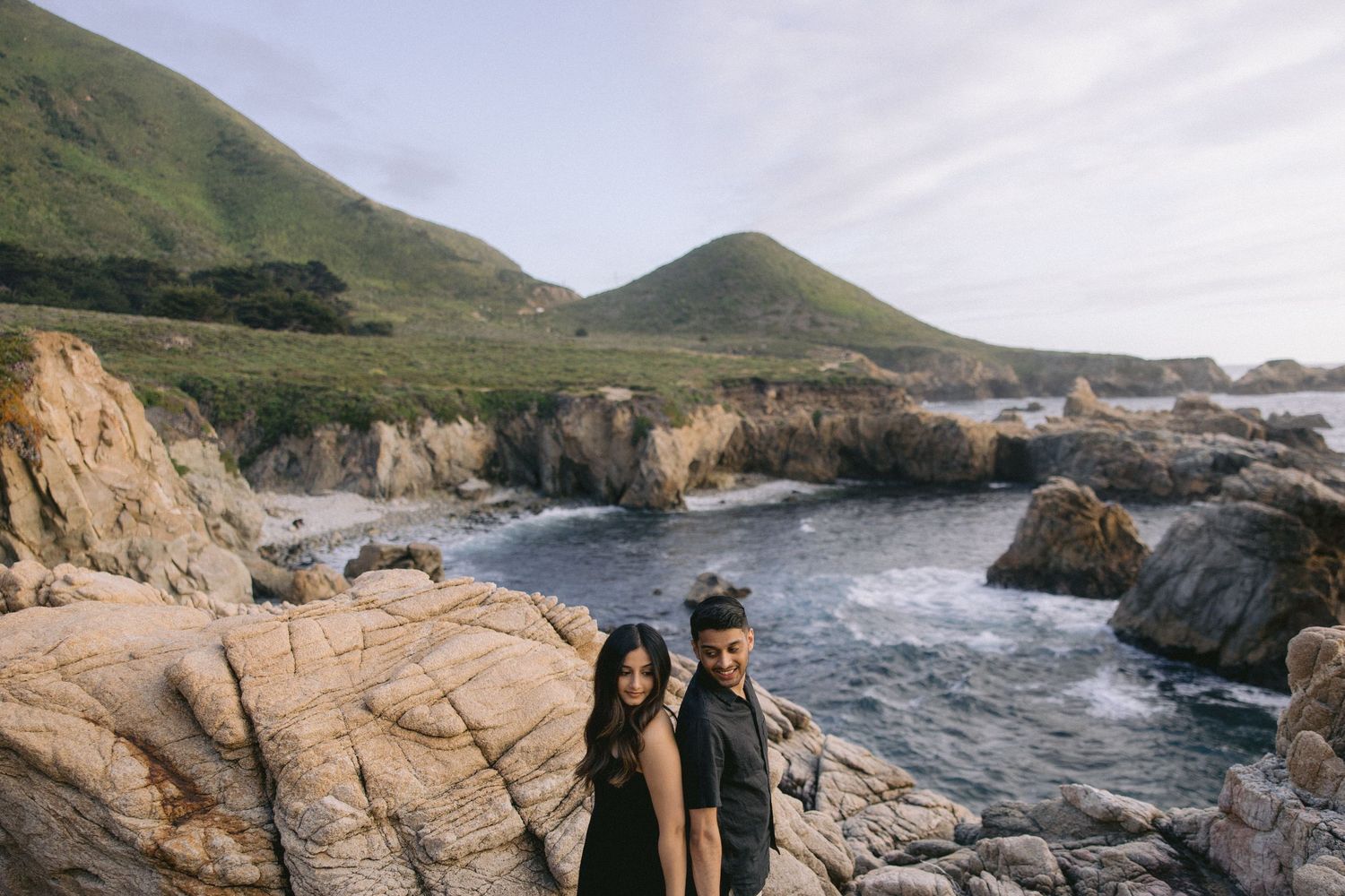 Two people stand together on rocky cliffs overlooking dramatic coastline at Big Sur, California.