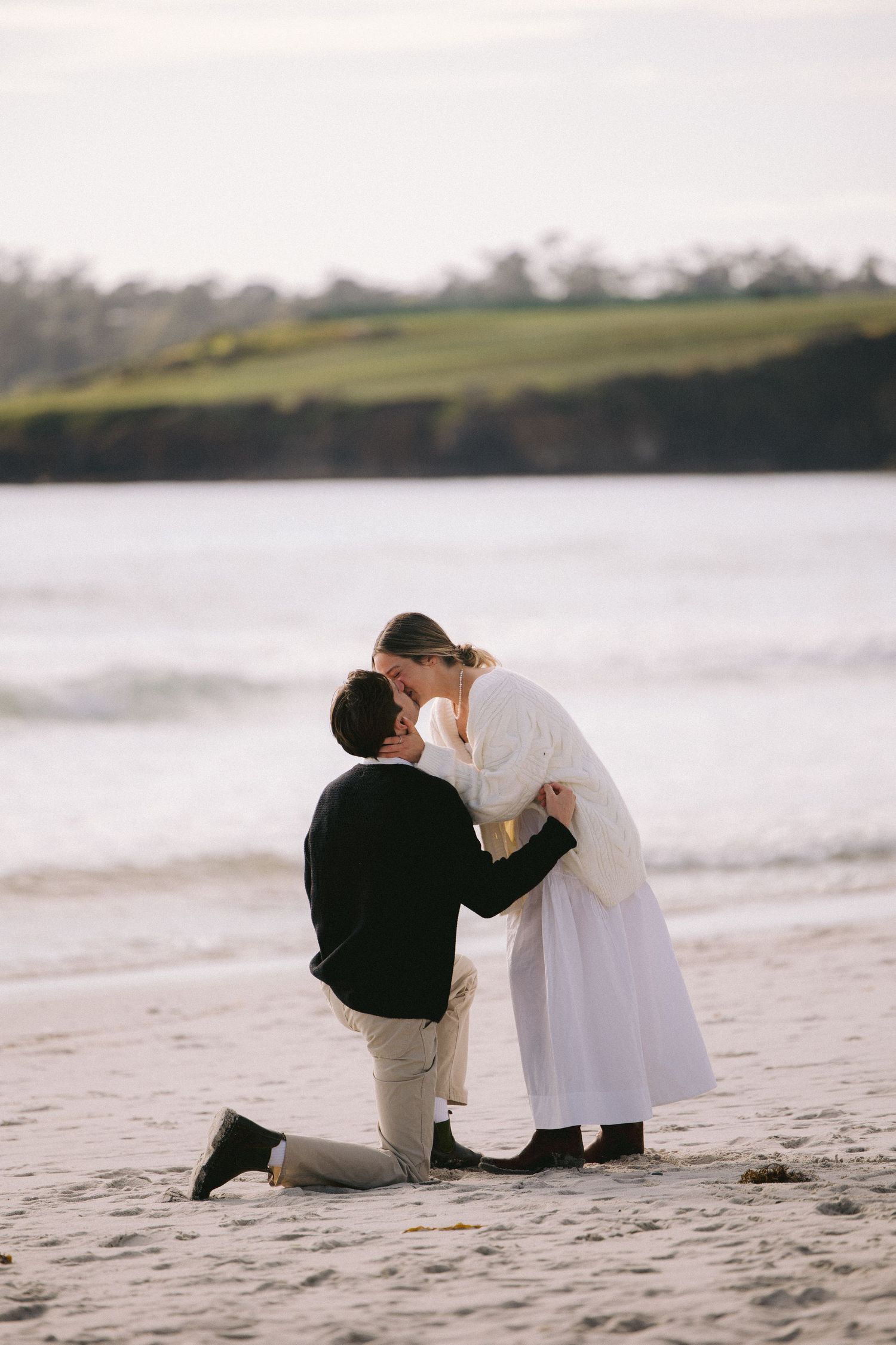A romantic couple embraces on a beach as one kneels down in the sand during sunset with rolling hills in the background.