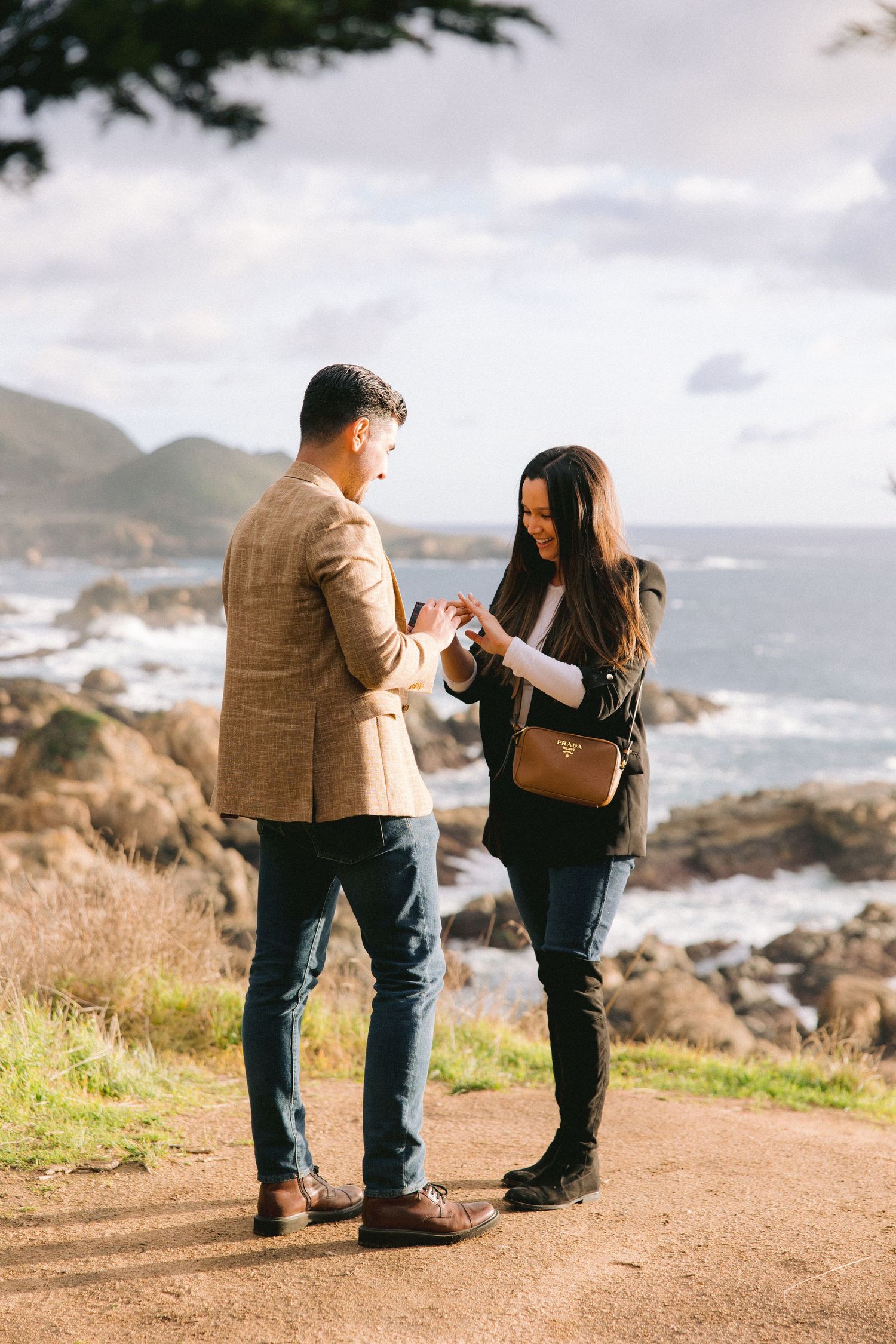 Couple shares an intimate proposal moment on a scenic coastal cliff overlooking the ocean and rocky shoreline.