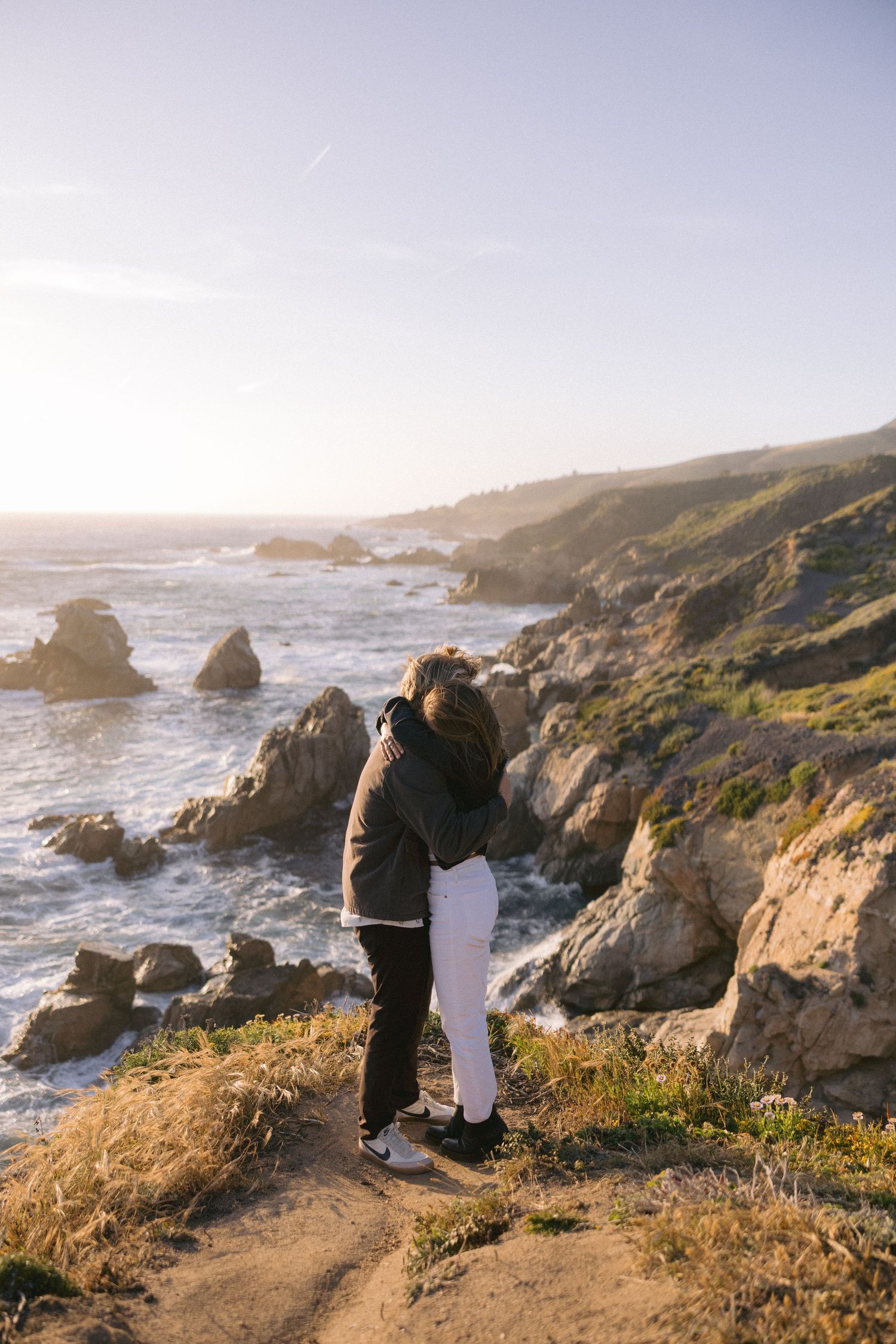 A couple embraces while standing on rocky coastal cliffs overlooking the ocean during golden hour at sunset.