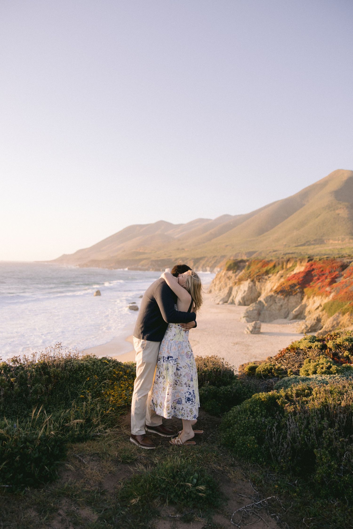 A romantic couple embraces on a cliff overlooking the dramatic Big Sur coastline at sunset in California.