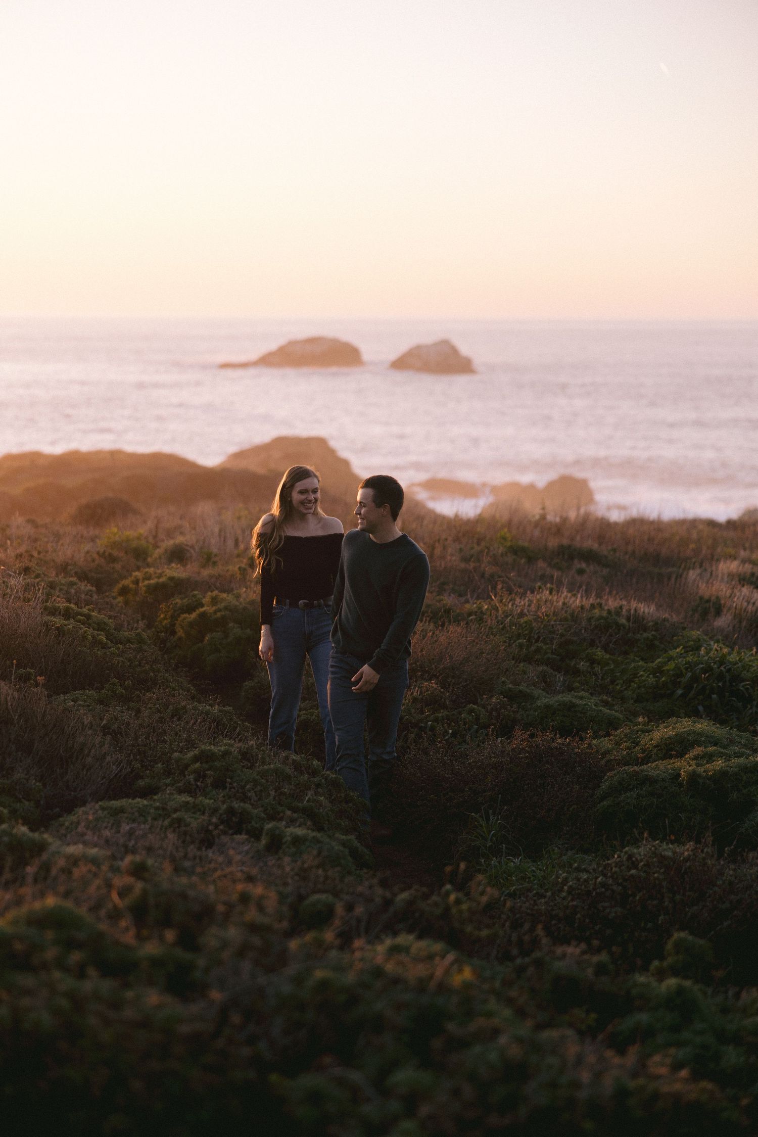 A couple stands on a coastal cliff at sunset with rocky islands visible in the misty ocean background.