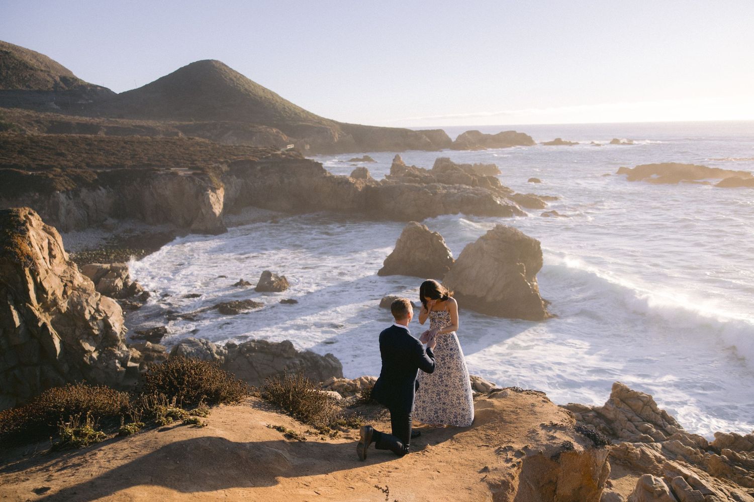 Two people stand on rocky cliff overlooking dramatic California coastline with crashing waves at sunset.