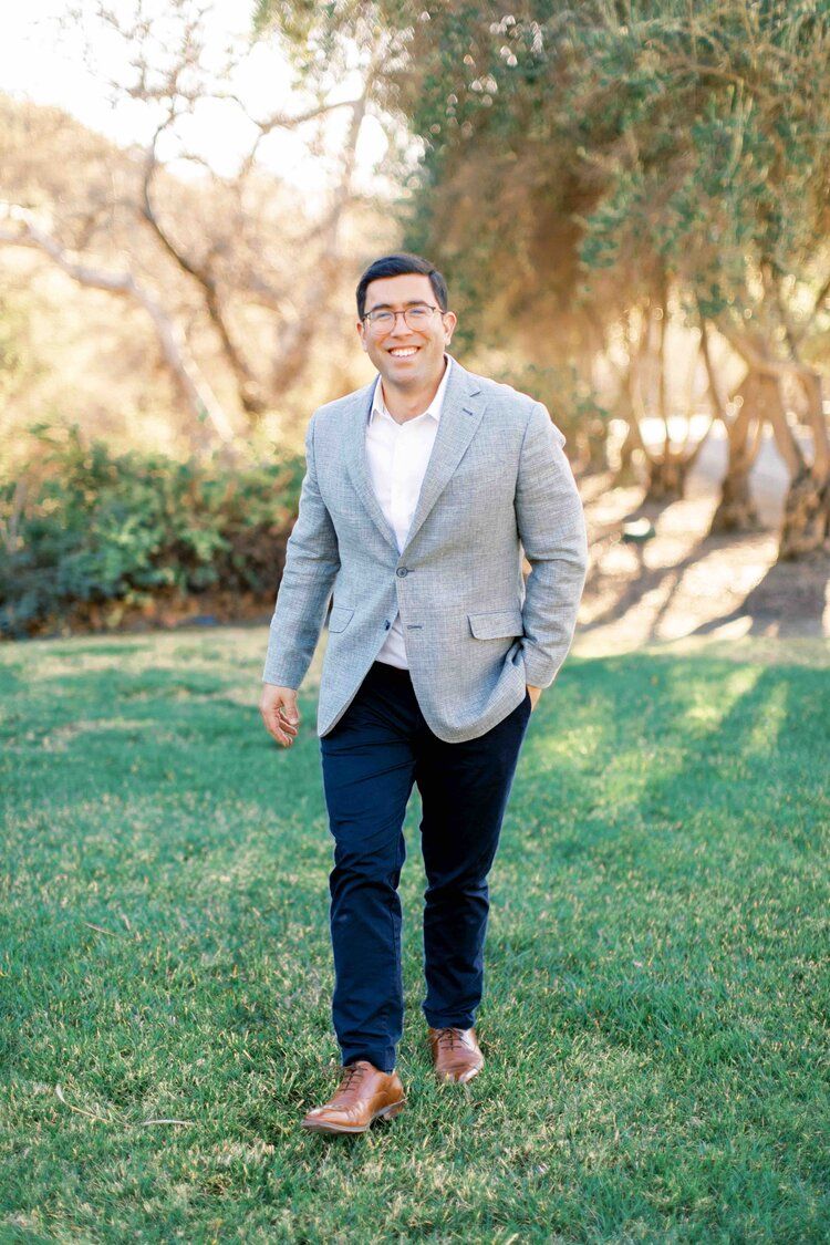 Professional outdoor portrait shows a person in a light gray blazer and navy pants standing on grass with trees in background.