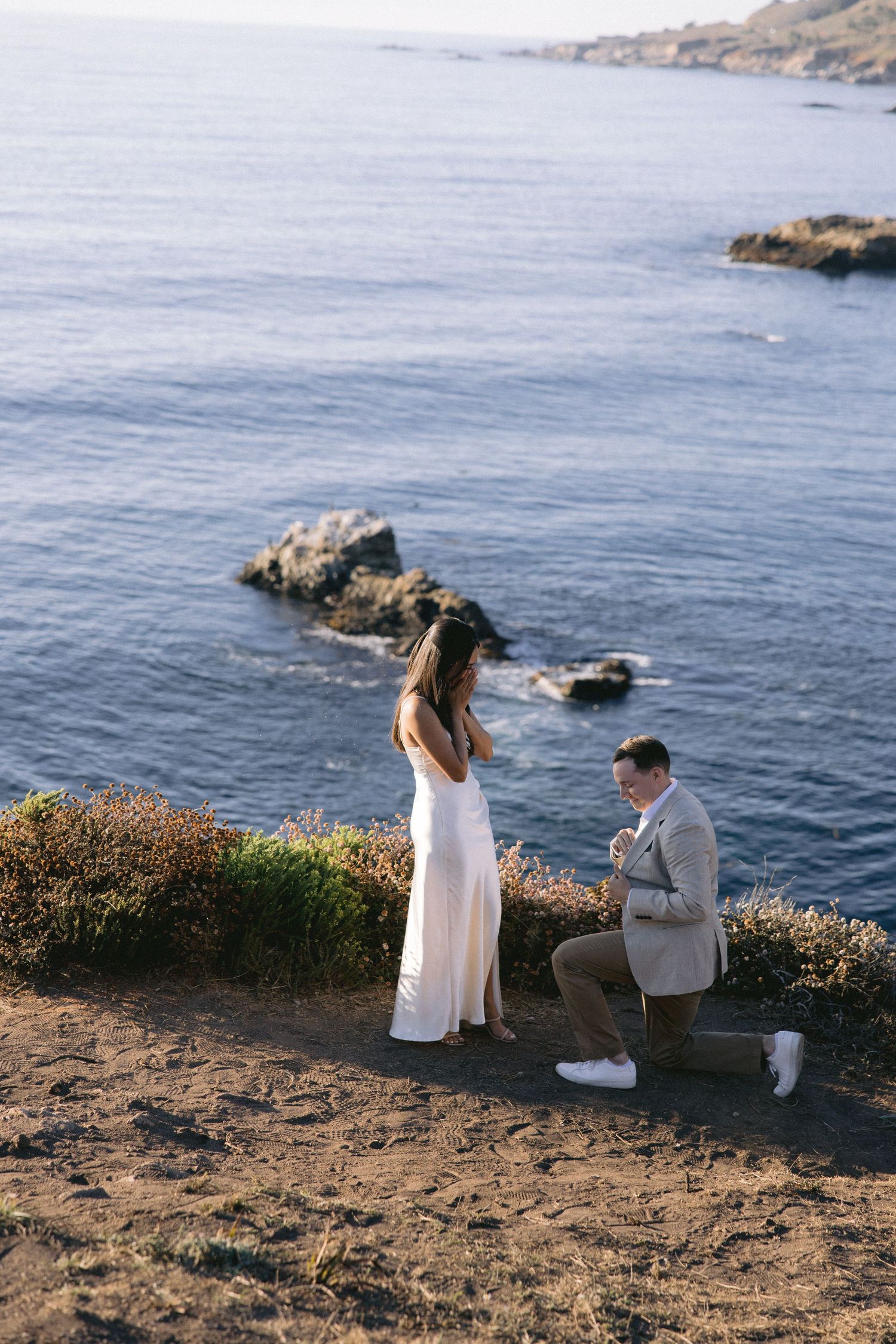 A romantic proposal moment on a scenic coastal cliff overlooking rocky outcrops and blue ocean waters at sunset.