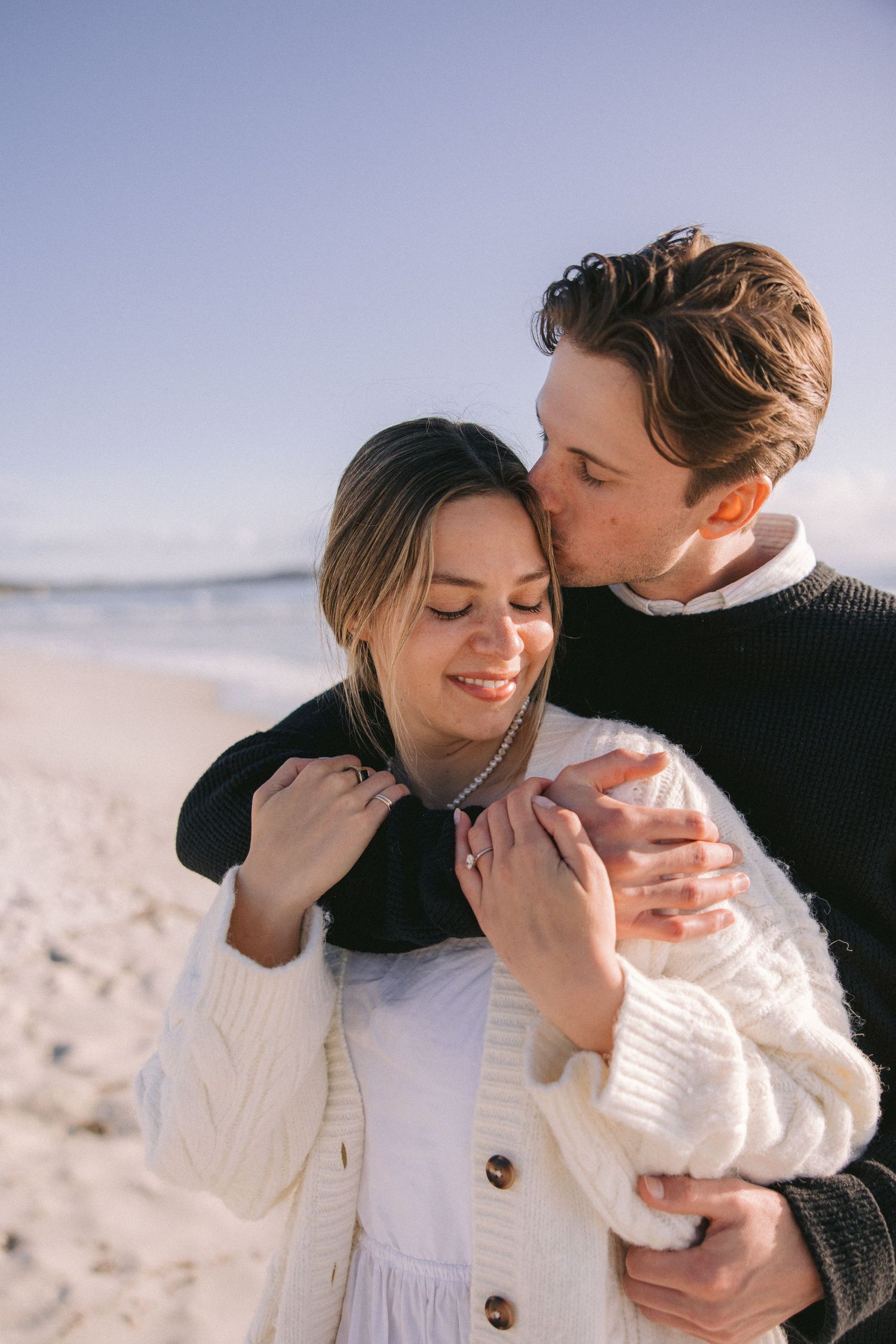 A loving couple shares an intimate embrace on a sunny beach with a cozy sweater and white coat.