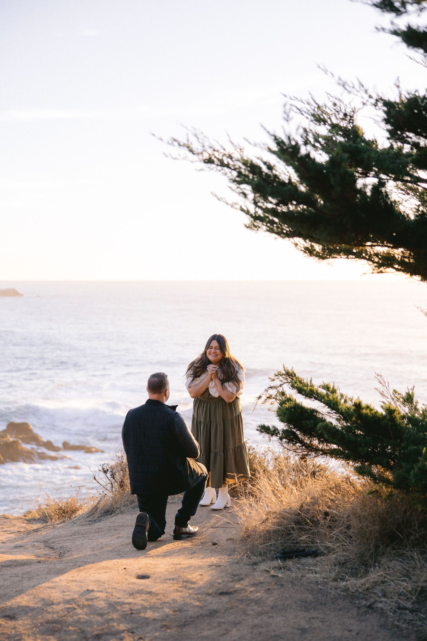 A romantic seaside proposal scene at sunset with cypress trees and ocean waves in the background.