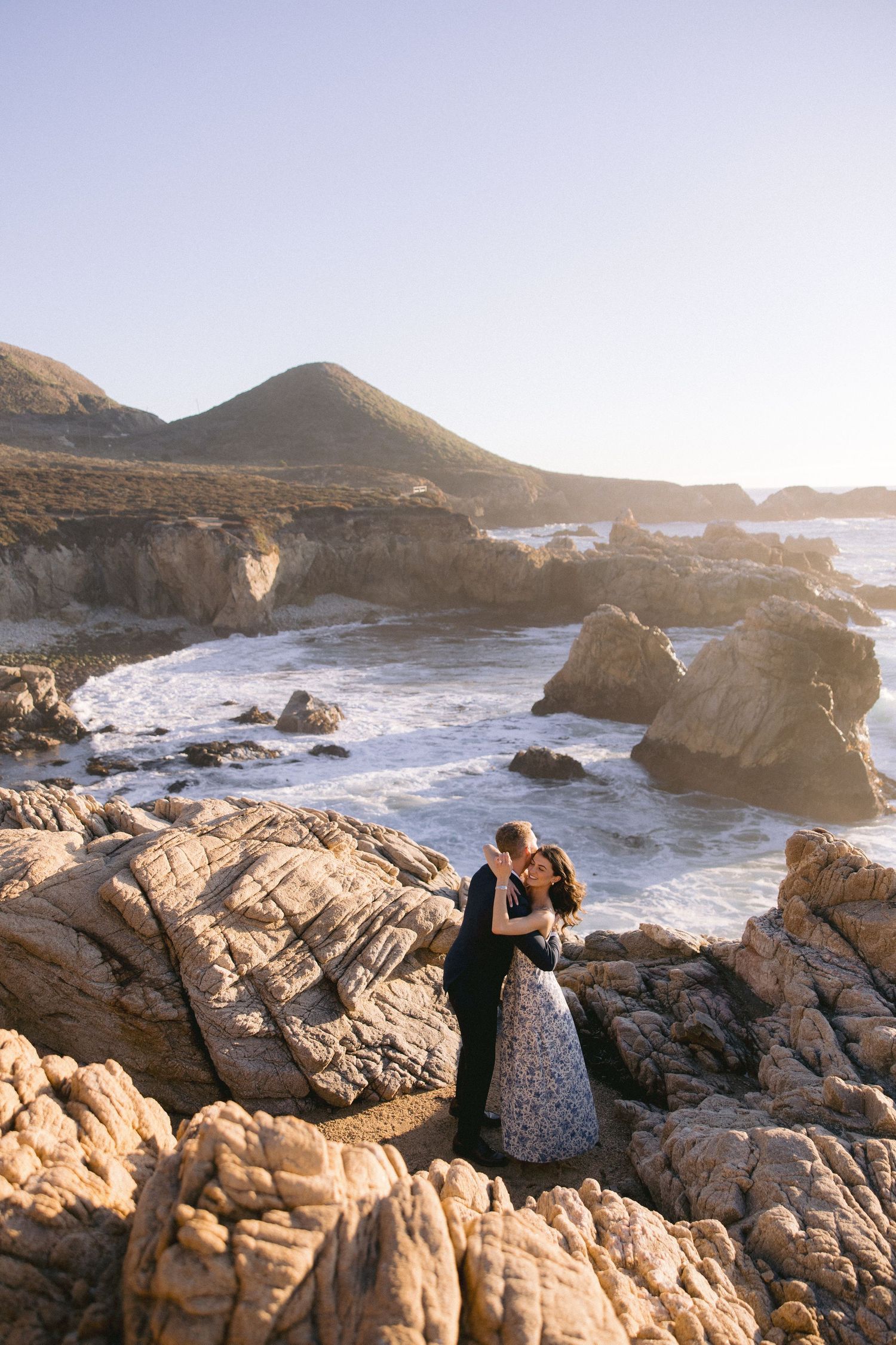 A couple embraces on rocky coastal cliffs at sunset with crashing waves and golden light illuminating the dramatic scene.