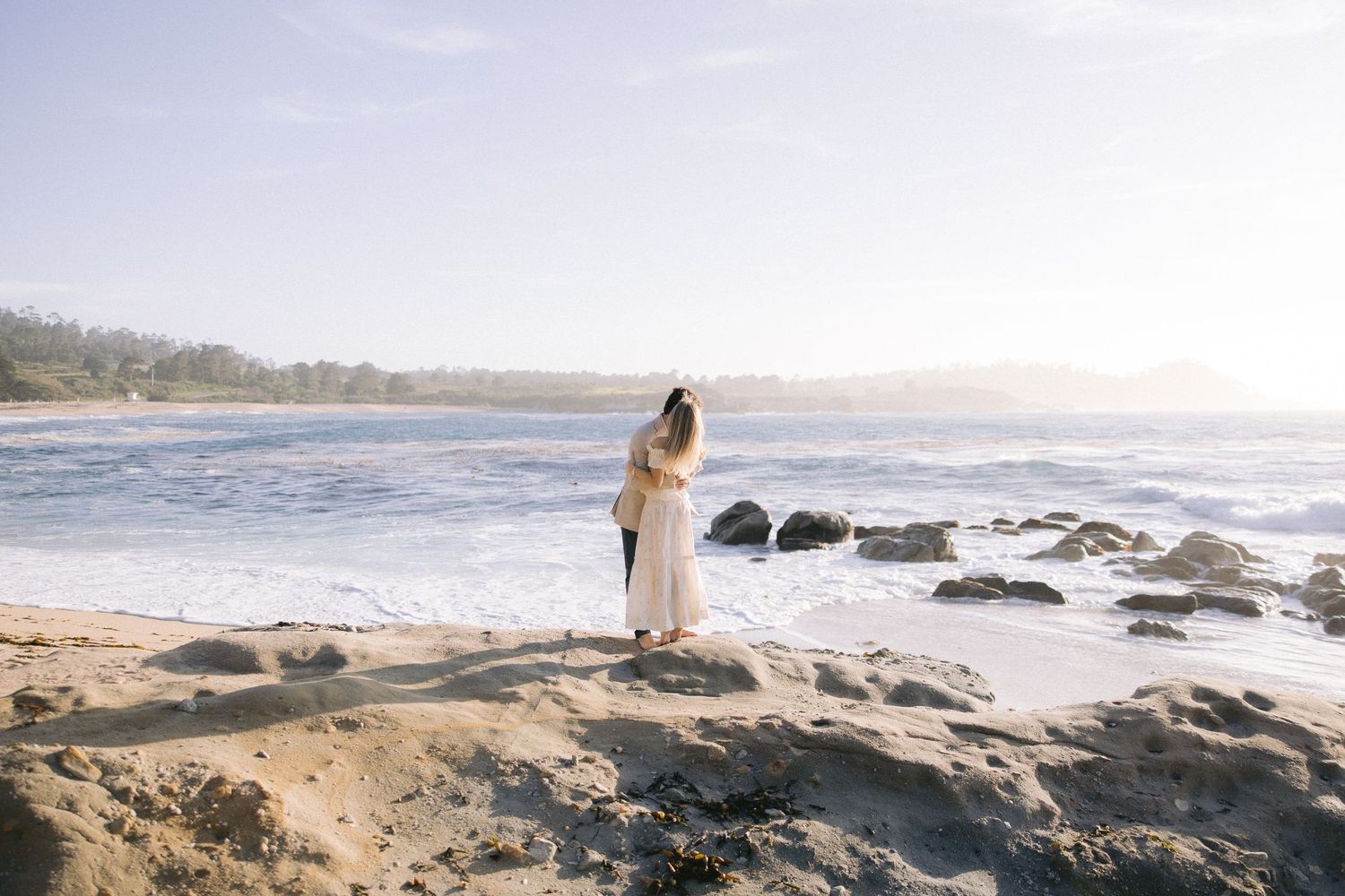 A person in white dress stands contemplatively on rocky coastal shoreline at sunset overlooking the ocean waves.