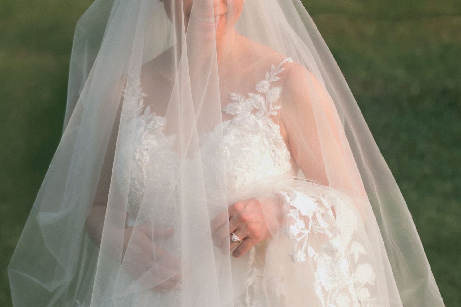 Bride in lace wedding dress with floral appliques covered by sheer veil outdoors on green grass background.