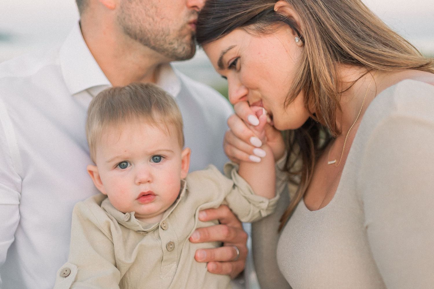 Tender family moment with baby in neutral tones against soft sky background.