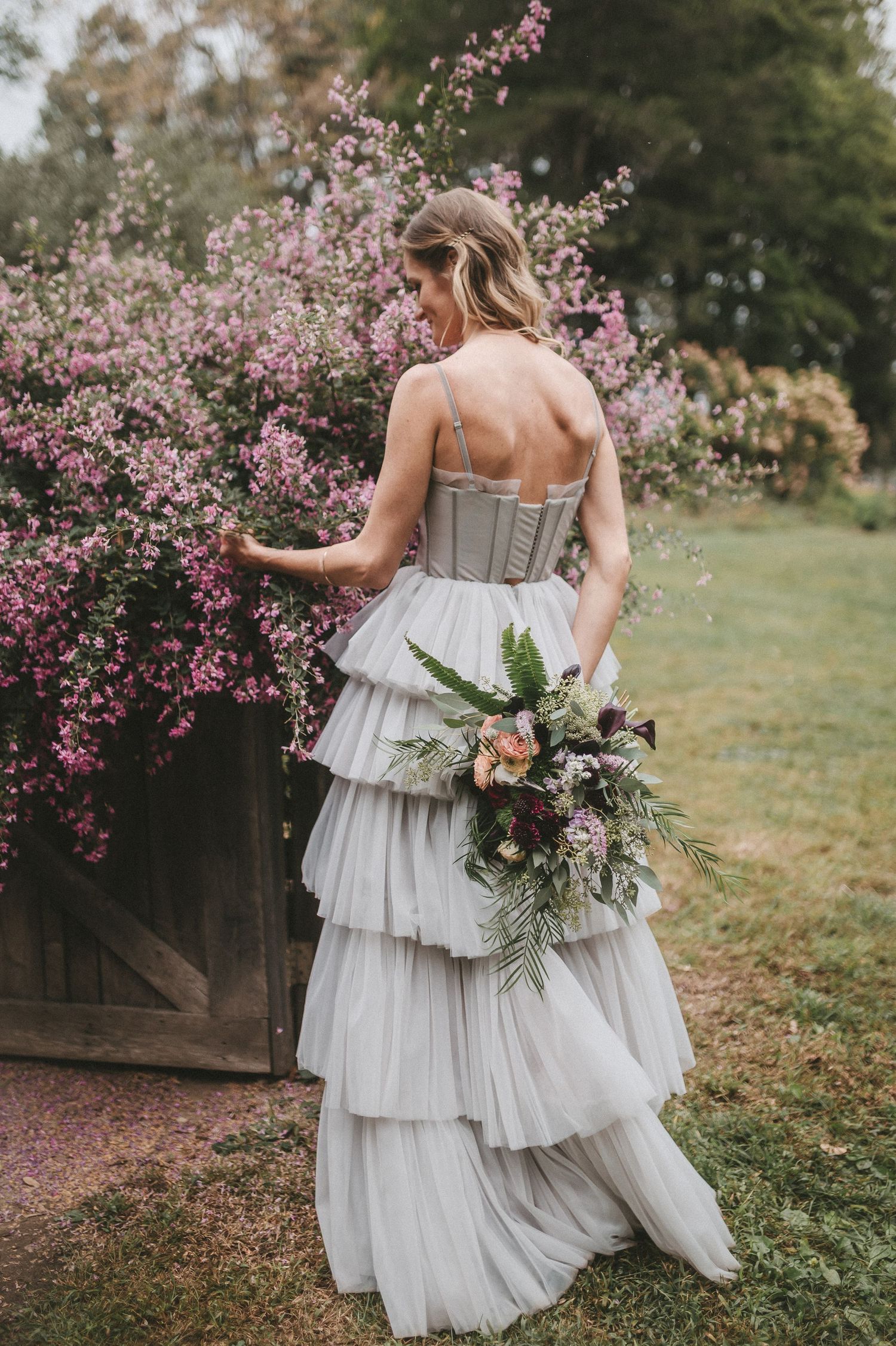 Bride in tiered white wedding gown with strapless corset bodice holds lush bouquet near blooming pink spring flowers.