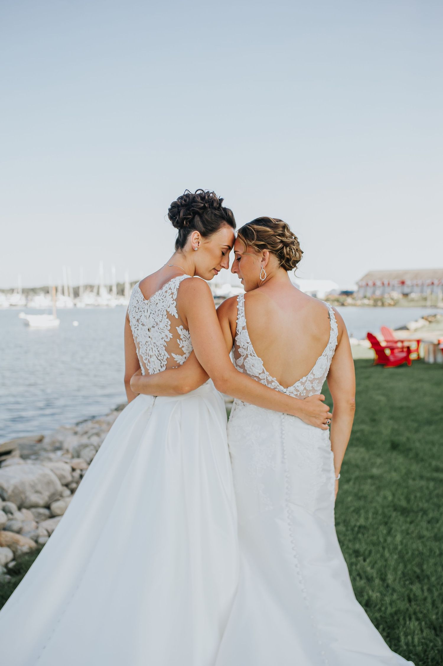Two brides in white wedding gowns embrace while overlooking a marina at sunset.