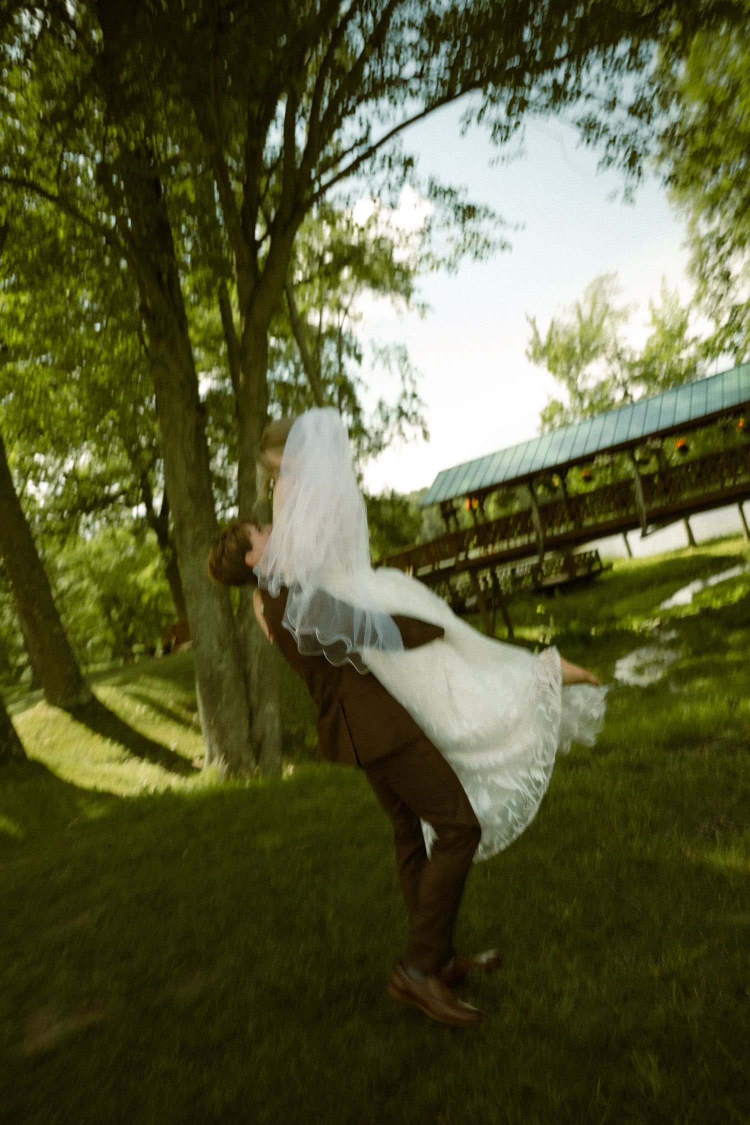 Groom lifting bride in white wedding dress and veil outdoors near trees on lush green lawn.