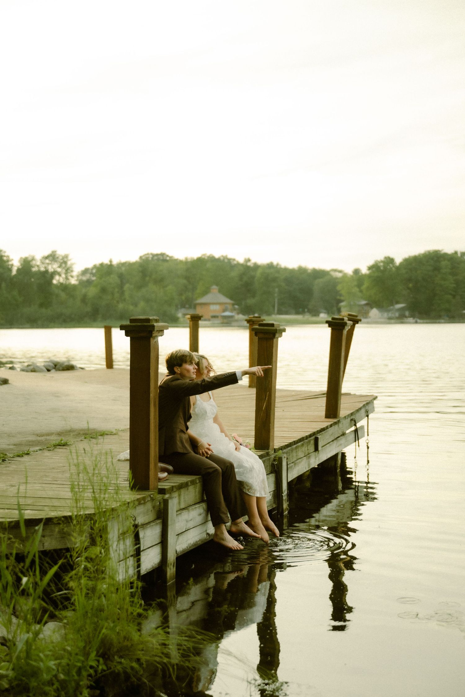 Couple sitting on a wooden dock by a calm lake at sunset, sharing a romantic moment with trees in the background.