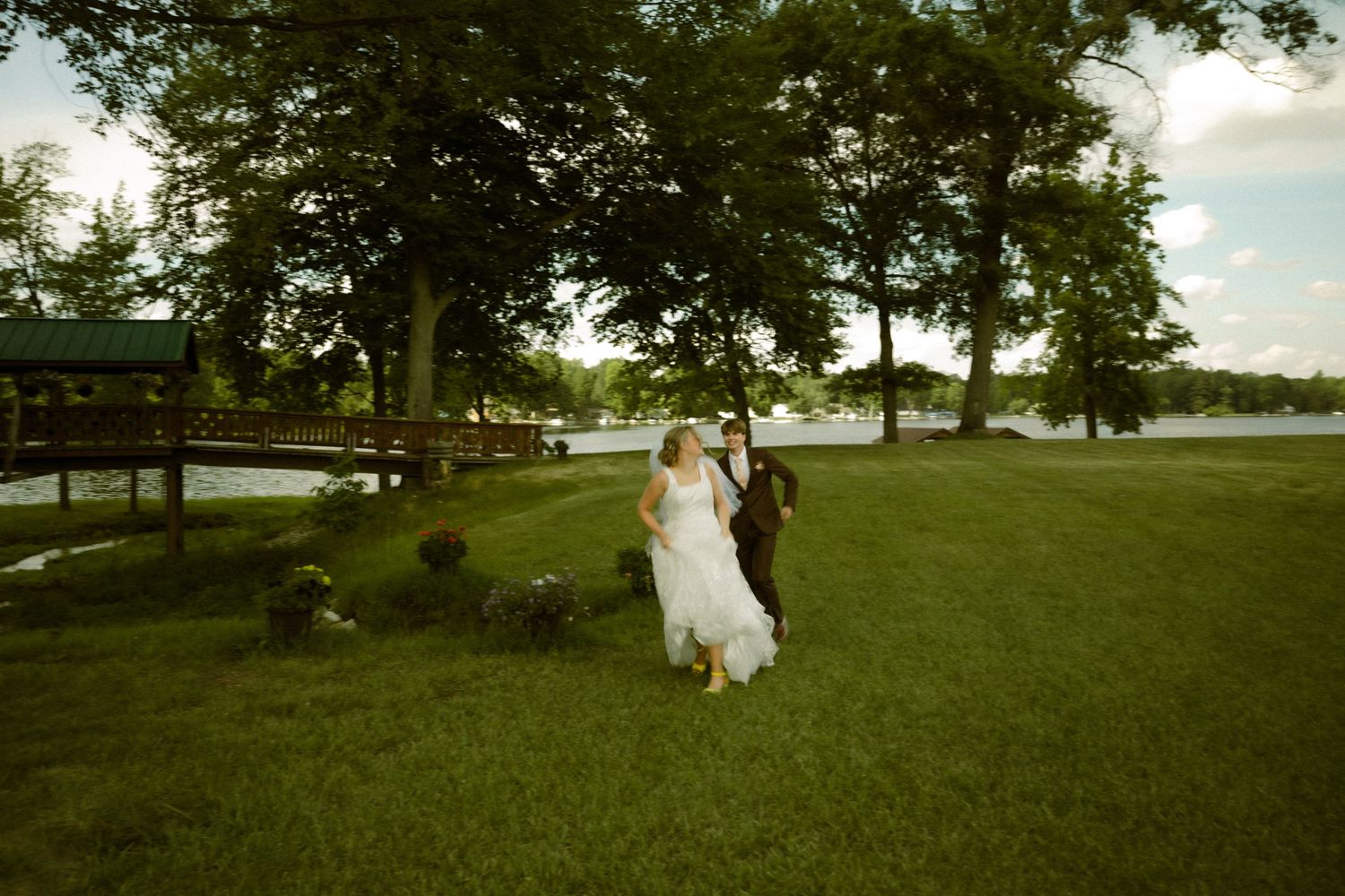 A bride and groom walk hand in hand across a lush green lawn near a lakeside venue surrounded by tall trees.