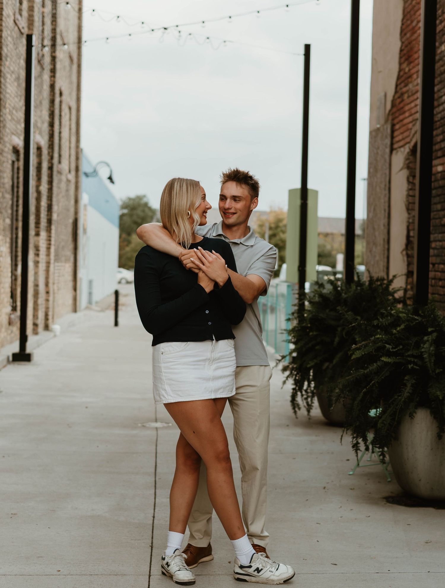 A couple shares an intimate moment together on a city sidewalk with string lights overhead.