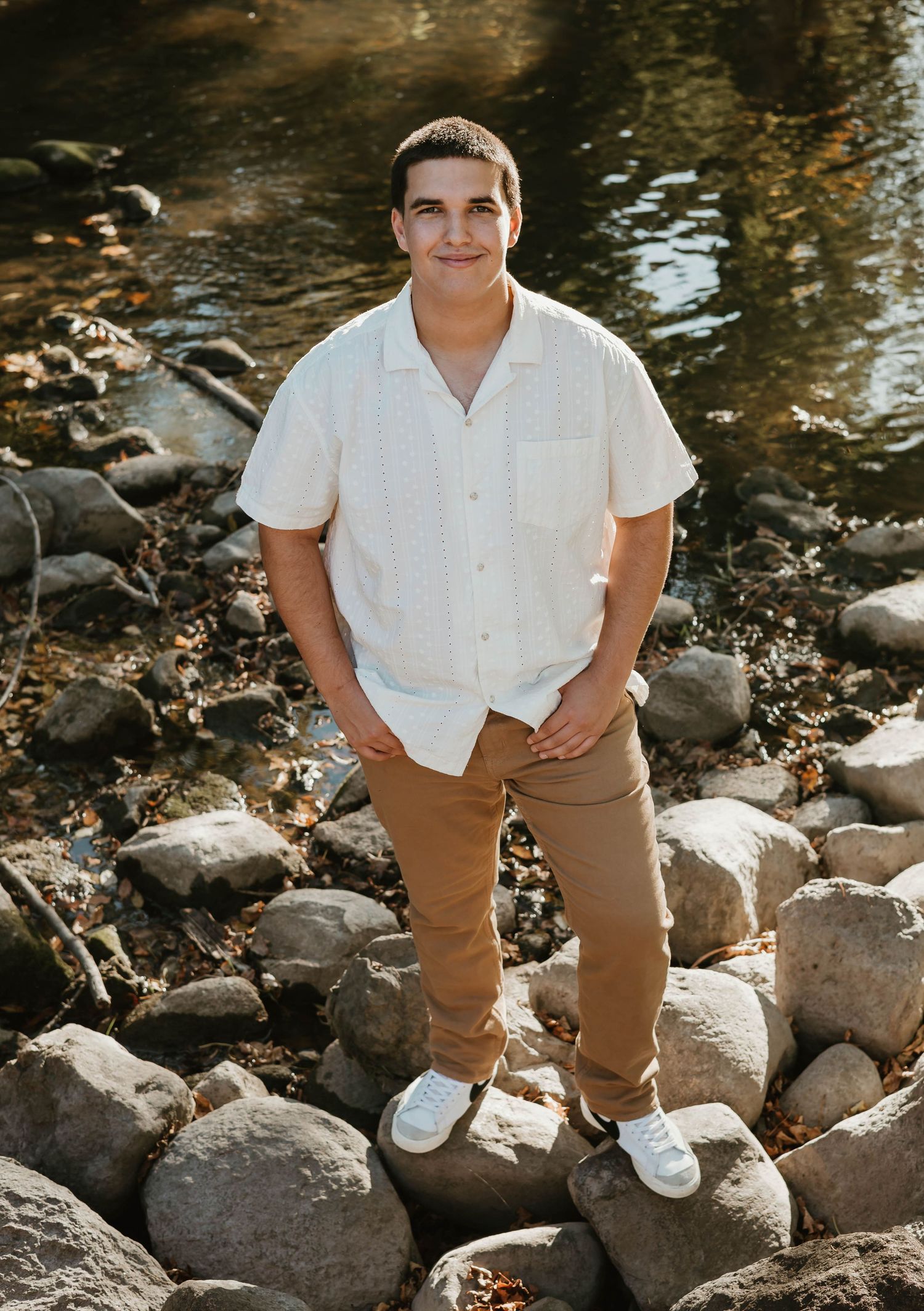 Young man in white shirt and tan pants standing on rocks by a river, smiling outdoors.