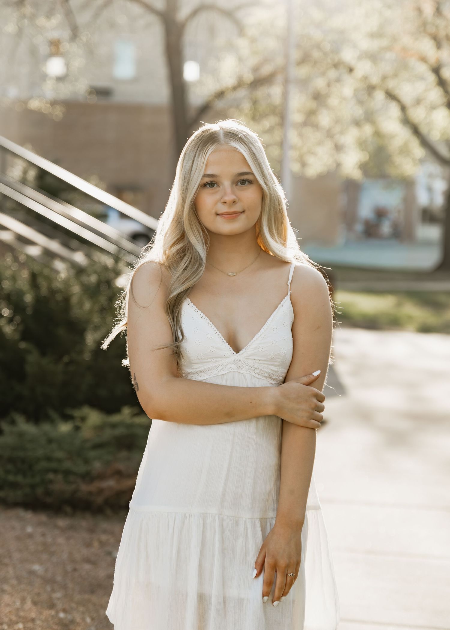 Young woman in white sundress posing outdoors in golden hour light with blurred urban background.