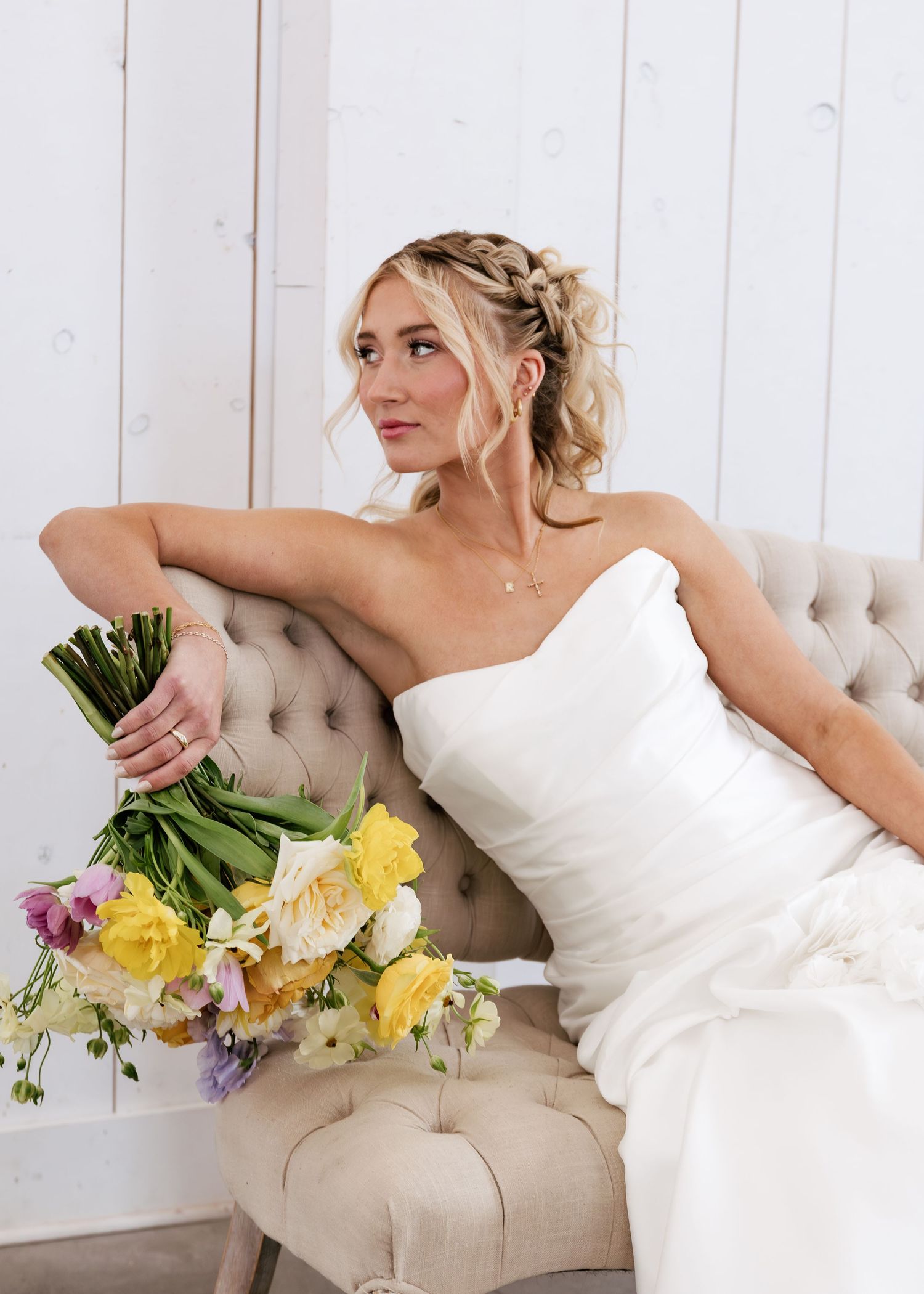 Bride in white strapless gown sits on tufted beige sofa holding colorful spring bouquet with yellow and purple flowers.