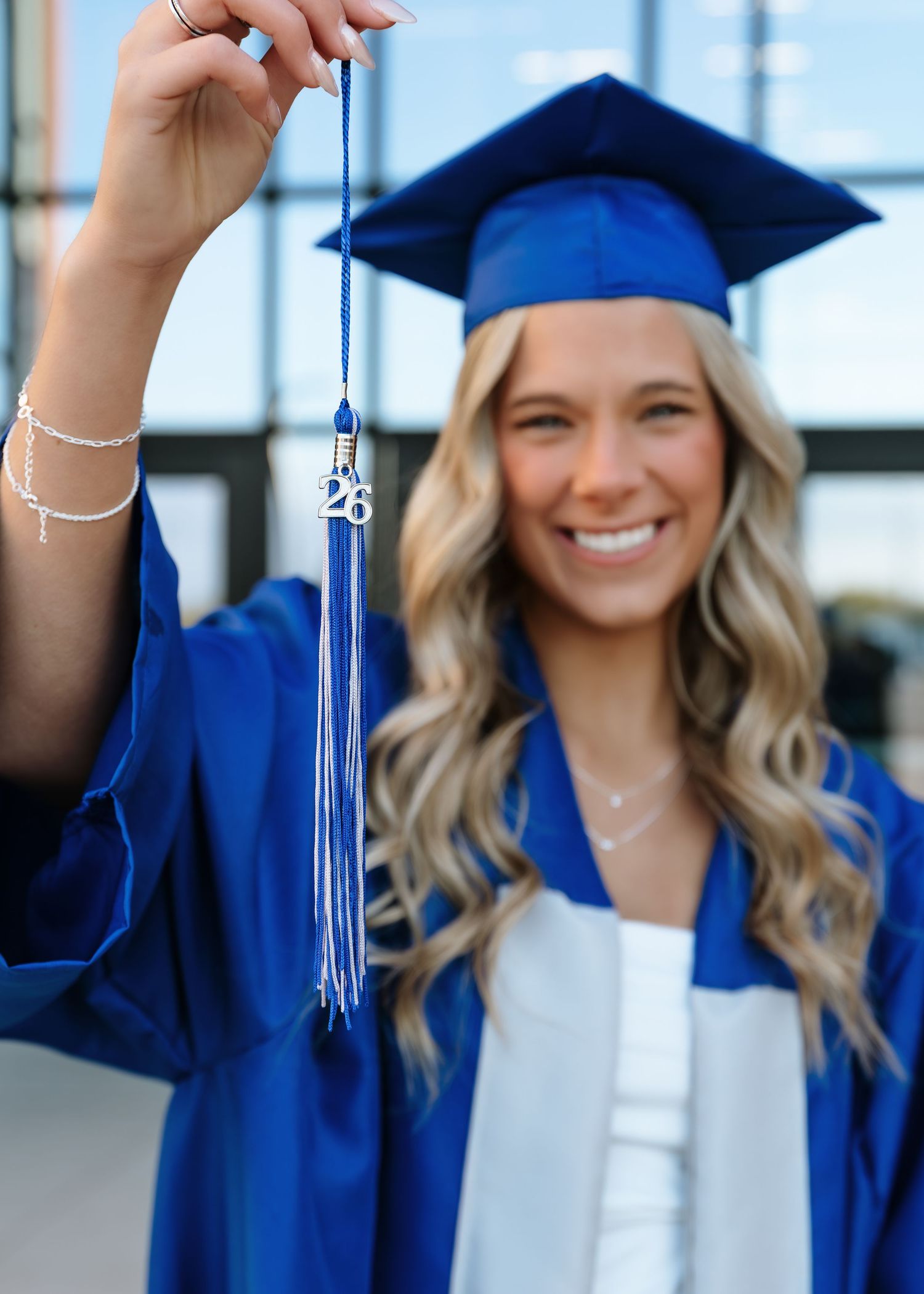 Smiling graduate in blue cap and gown holds tassel with 26 charm, celebrating class of 2026 graduation outdoors.