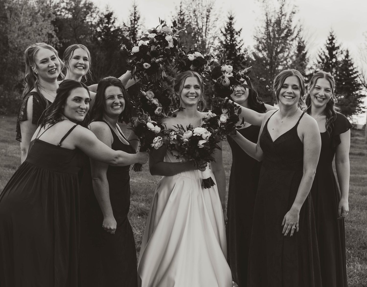 Wedding party in black dresses poses with the bride outdoors.