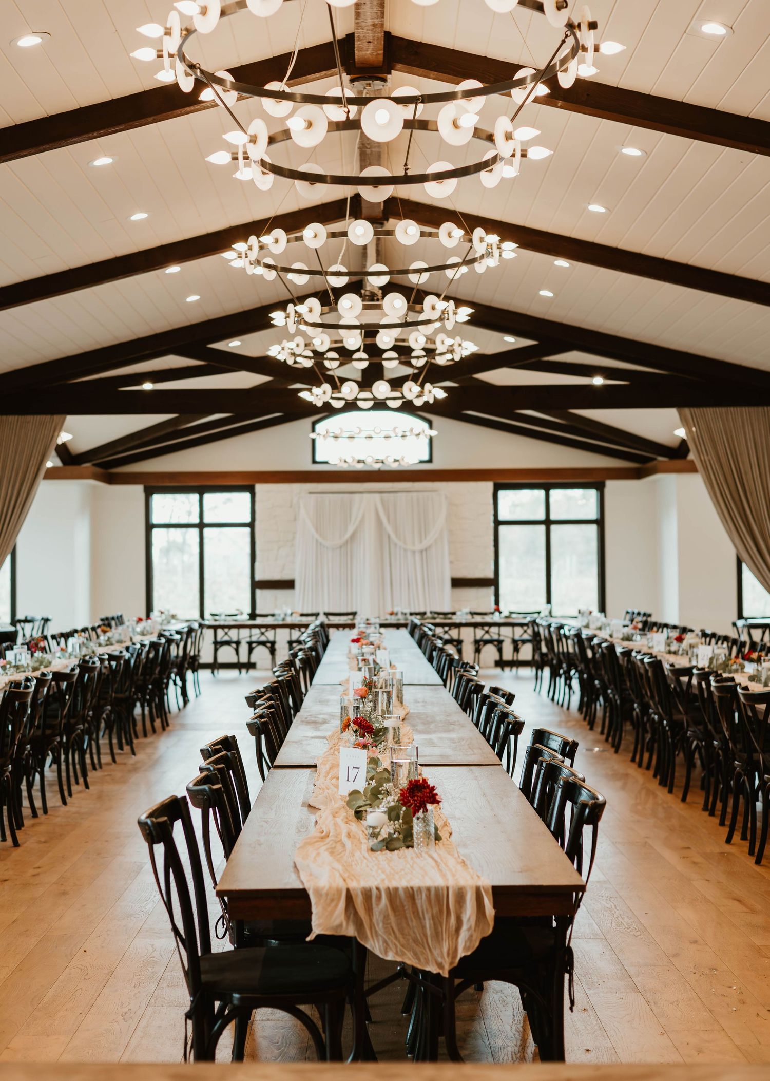 Elegant wedding reception hall with tiered chandeliers and long tables set beneath exposed wooden beams.
