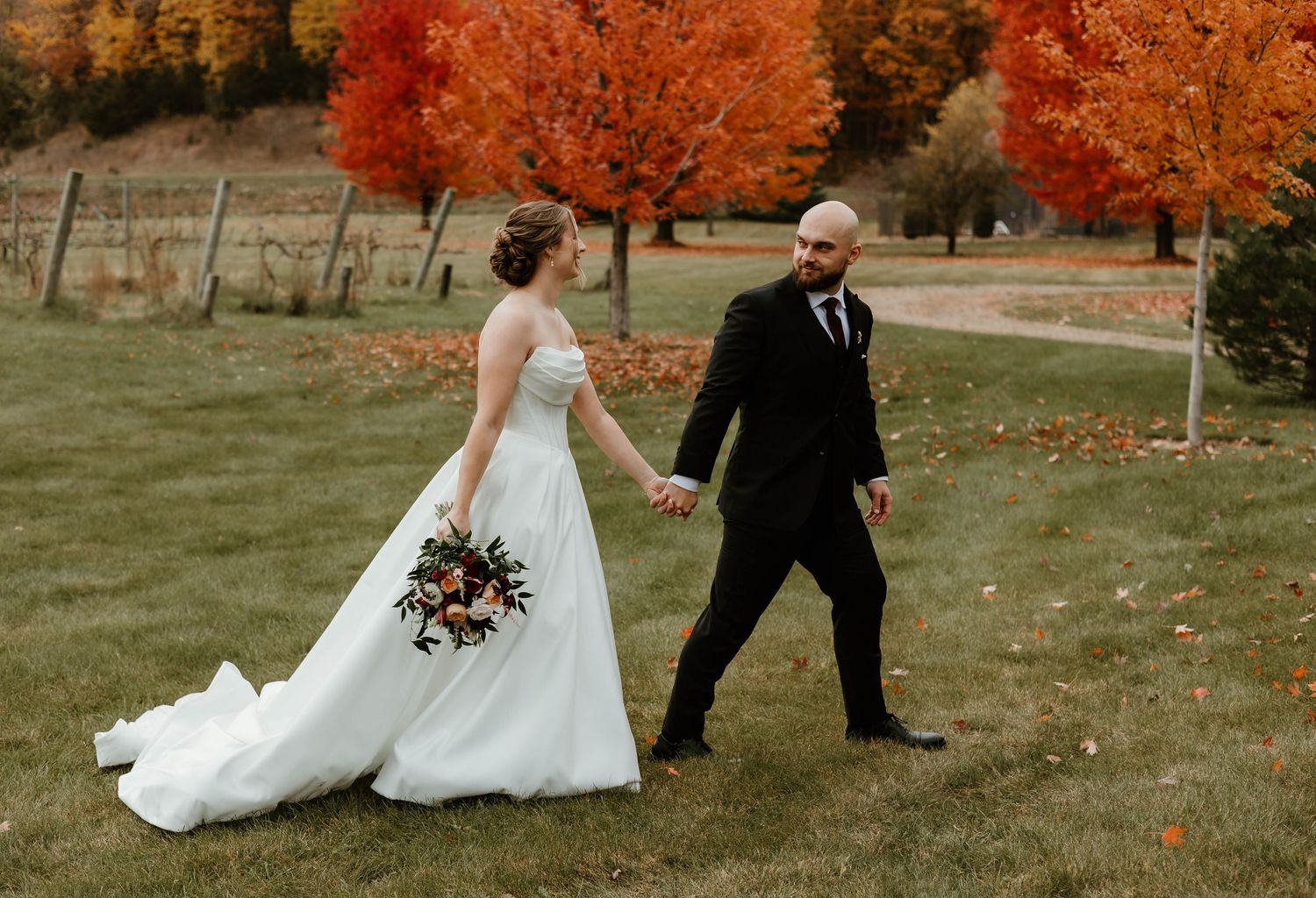 A couple walks through a scenic autumn landscape with vibrant orange trees and fallen leaves on a sunny day.