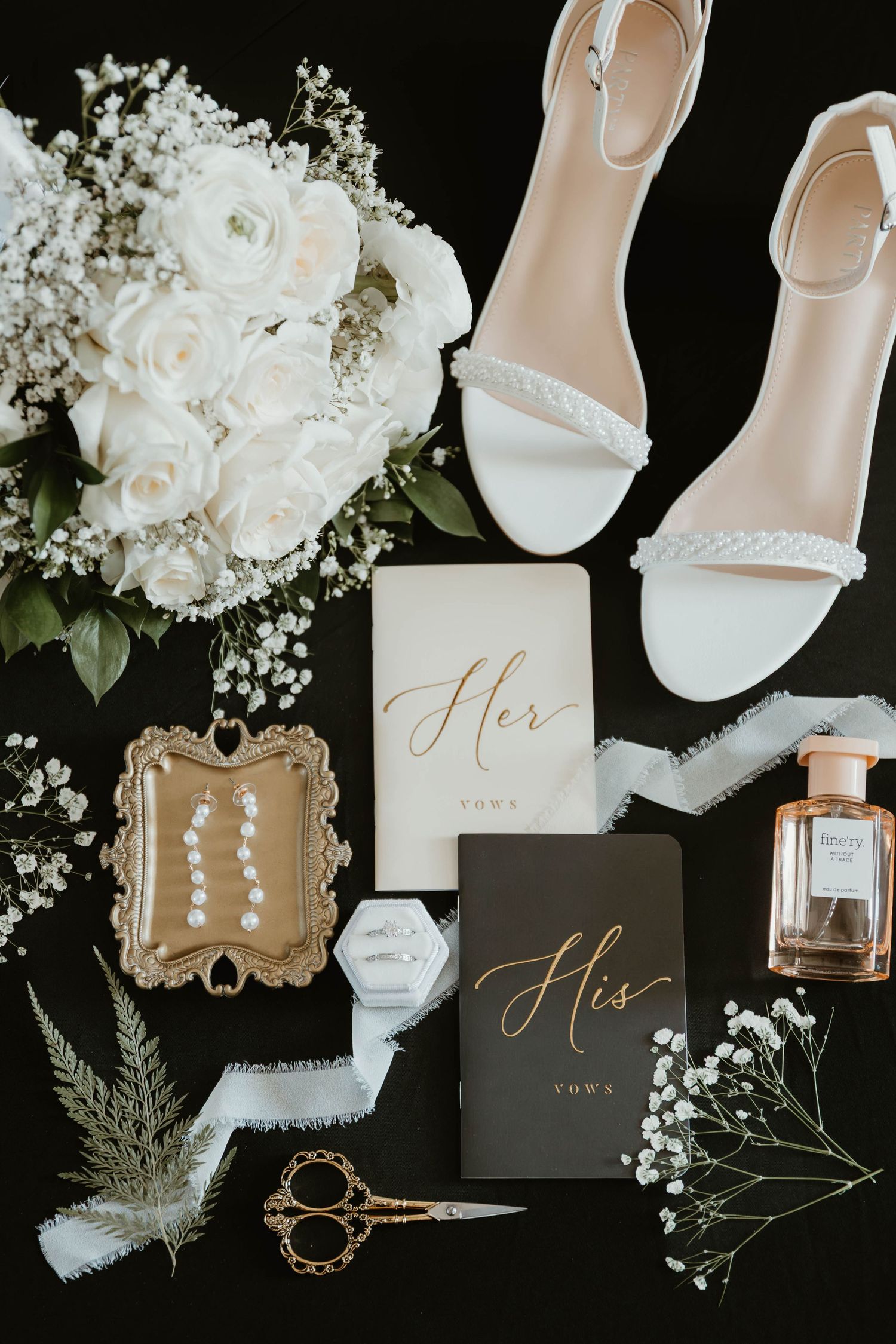 Wedding details flatlay with white roses, bridal shoes, vows books, perfume and delicate accessories on black background.