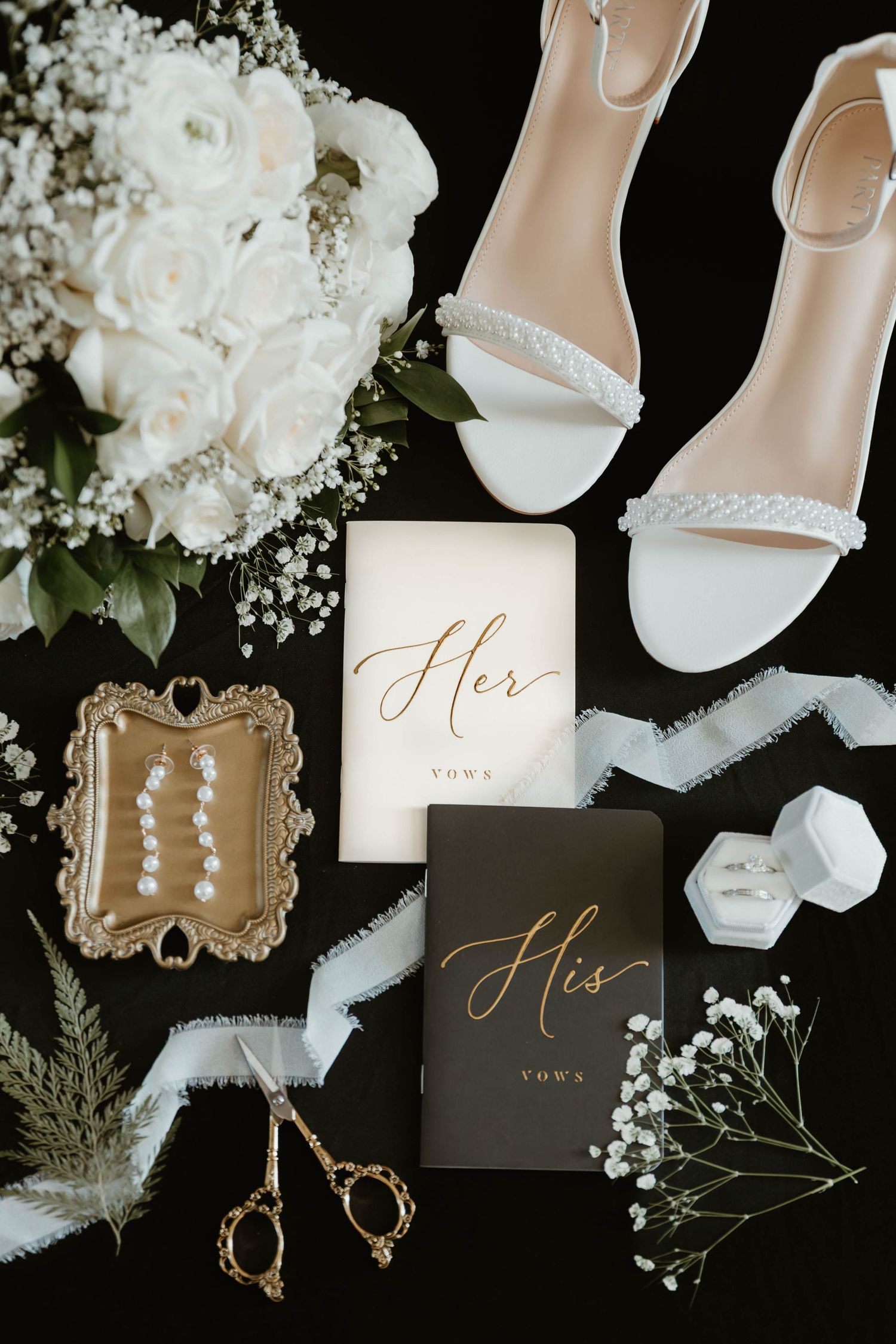 Elegant wedding details flatlay with white shoes, floral arrangements, jewelry and His & Her cards on black background.